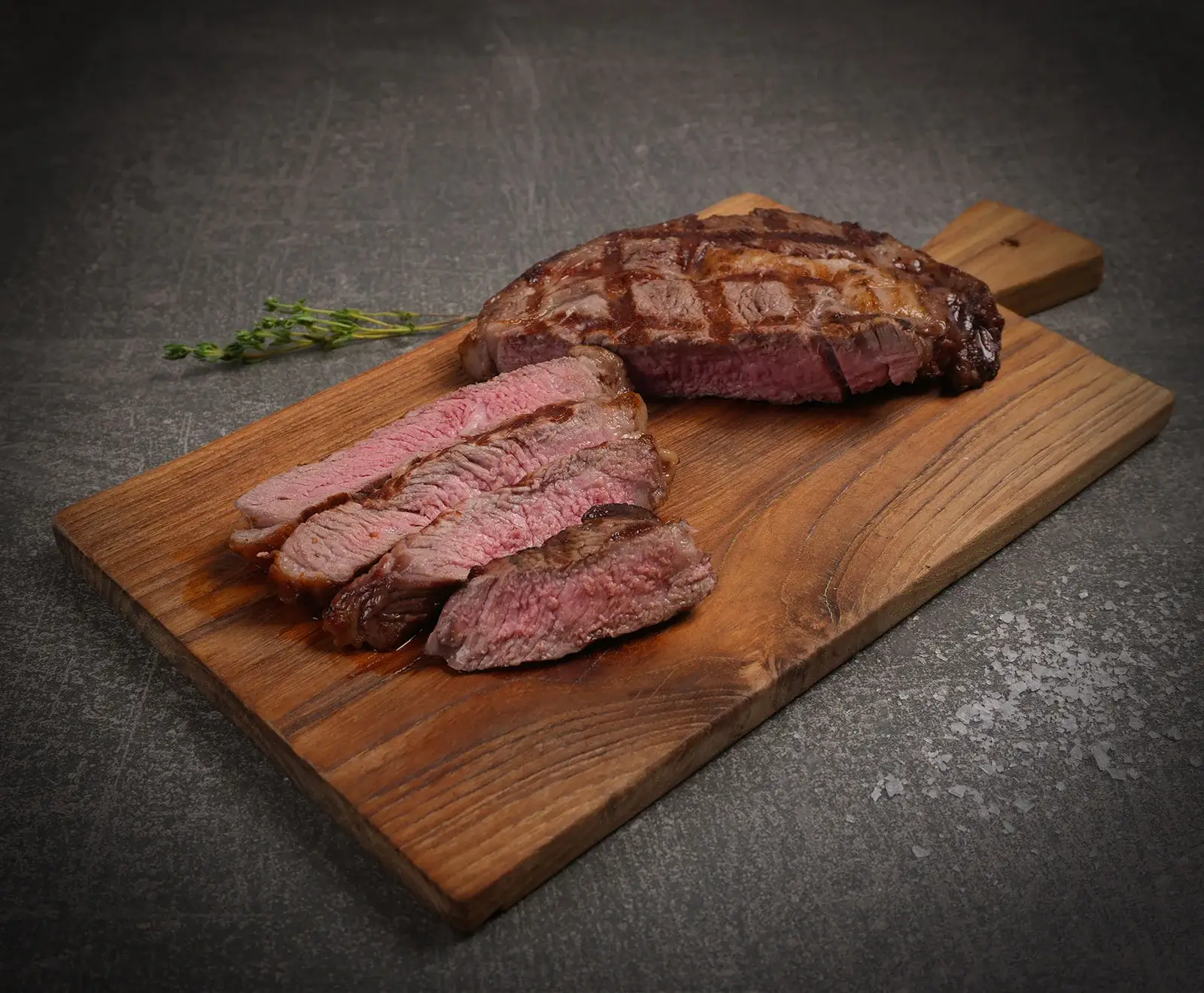 Ein saftiges, medium-rare gegrilltes Steak mit Grillstreifen, teilweise in Scheiben geschnitten, liegt auf einem Holzschneidebrett. Daneben sind frische Thymianzweige zu sehen.