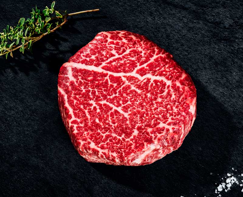 Ein rohes, stark marmoriertes Rindersteak (vermutlich Wagyu) liegt auf einer dunklen, strukturierten Oberfläche. Daneben sind ein frischer Thymianzweig und grobes Meersalz zu sehen. Das Fleisch ist leuchtend rot mit feinen weißen Fettadern.