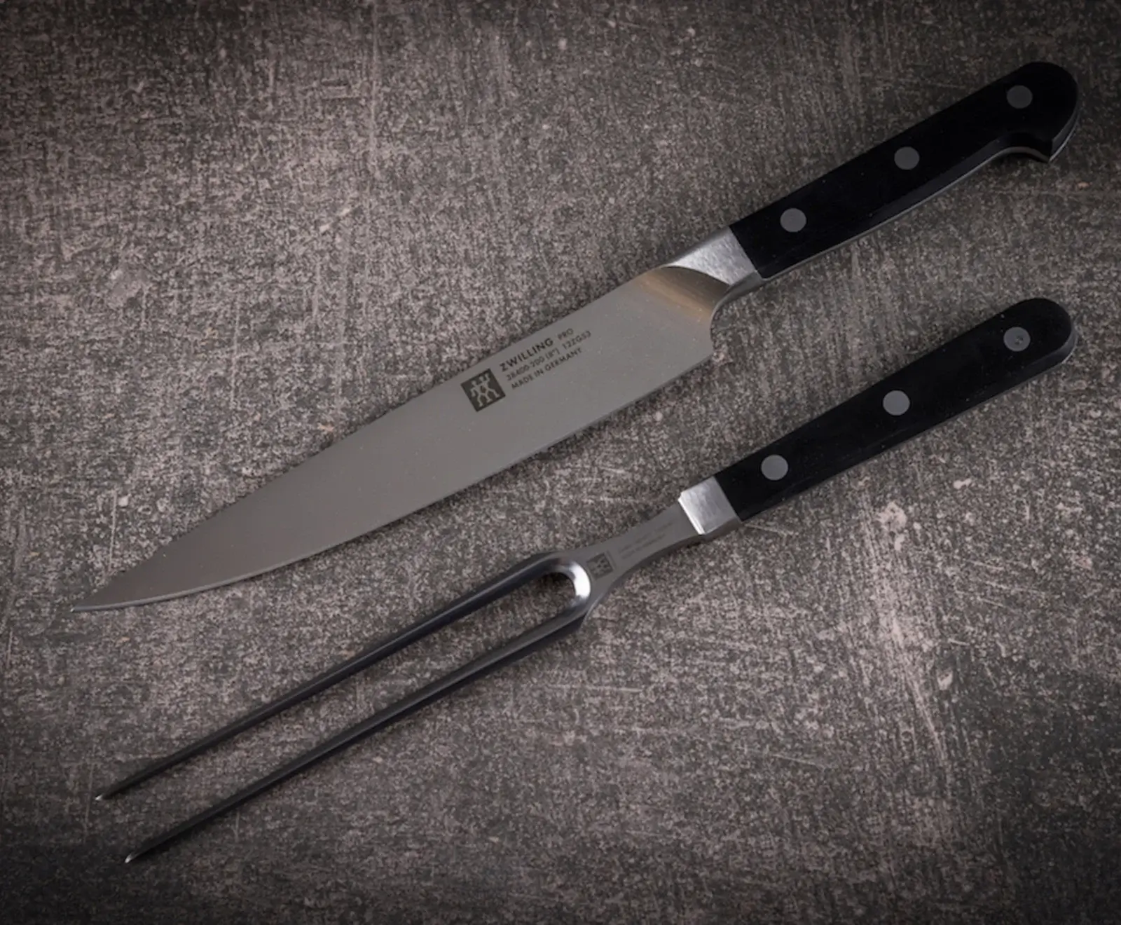 Ein Zwilling Pro Tranchiermesser und eine Tranchiergabel mit schwarzen Griffen und silbernen Klingen/Zinken liegen diagonal auf einer dunklen, strukturierten Oberfläche.