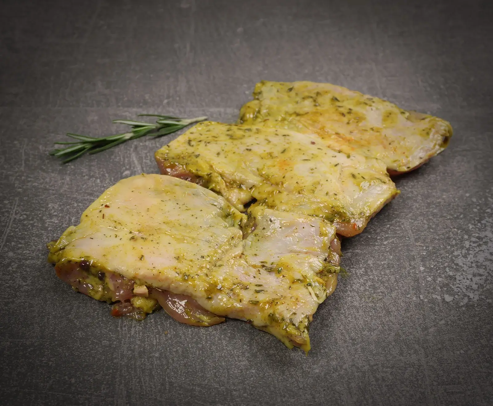Drei rohe, marinierte Hähnchenteile mit gelb-grüner Kräutermarinade und einem frischen Rosmarinzweig auf einem dunklen, strukturierten Hintergrund.