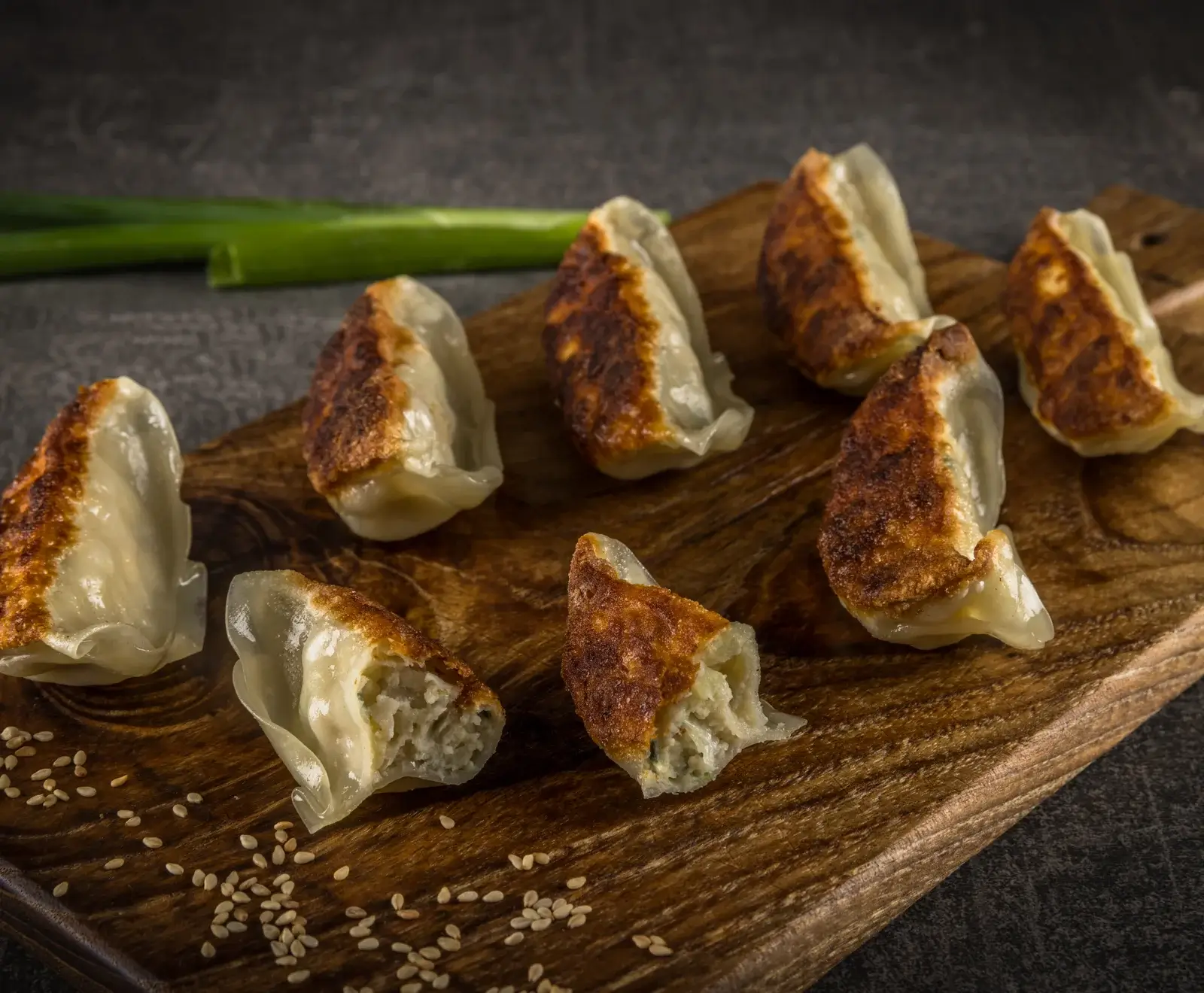 Acht goldbraun gebratene Teigtaschen (Gyoza) liegen auf einem dunklen Holzbrett, umgeben von Sesamkörnern. Eine Teigtasche ist aufgebrochen und zeigt die helle Füllung. Im Hintergrund liegt eine Lauchzwiebel.