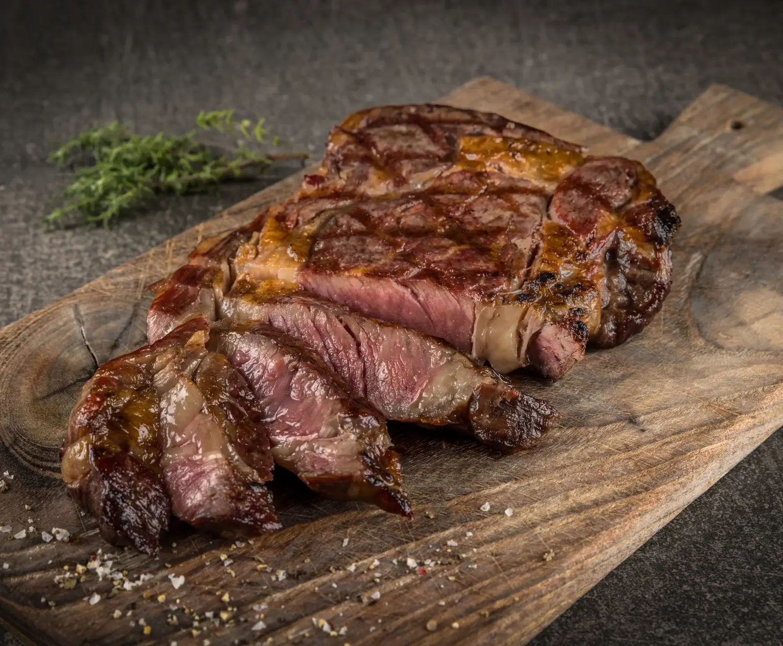 Ein saftiges, gegrilltes Rindersteak, teilweise in Scheiben geschnitten, liegt auf einem rustikalen Holzbrett. Es ist medium rare gegart und mit grobem Salz, Pfeffer und Thymian garniert.