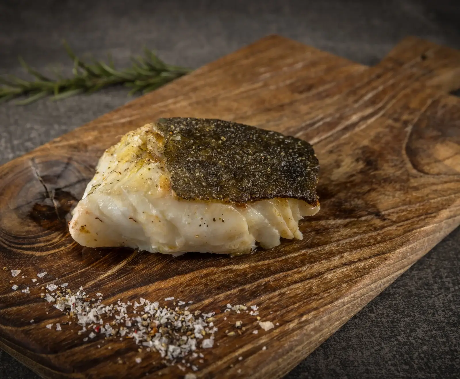 Nahaufnahme eines gebratenen Fischfilets mit knuspriger Haut auf einem rustikalen Holzbrett, daneben grobes Salz, Pfeffer und ein Rosmarinzweig.