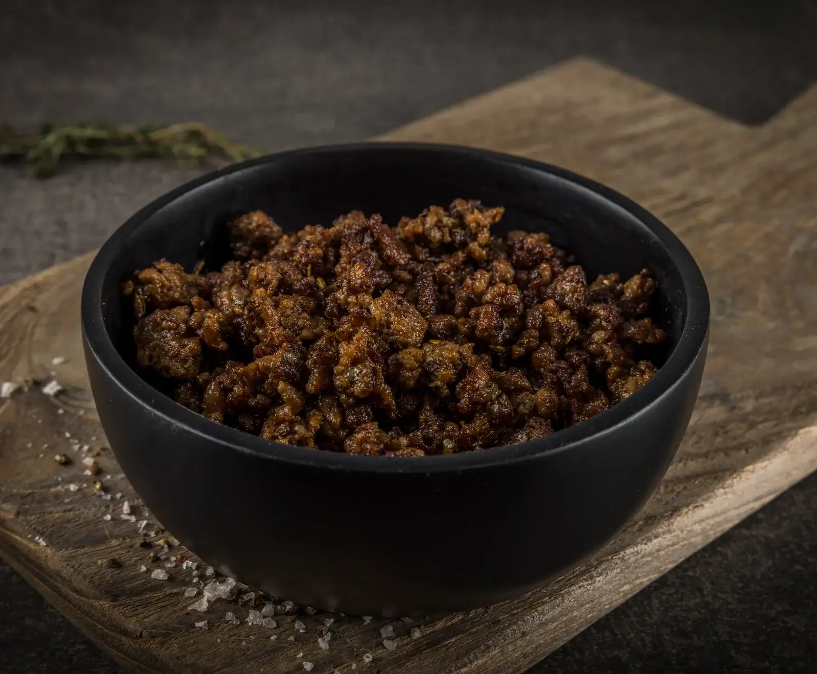 Schwarze Schale gefüllt mit braunem, gebratenem Gehackten (Fleisch oder pflanzlich) auf einem rustikalen Holzbrett. Grobes Salz und Pfeffer sind verstreut, grüne Kräuter im unscharfen Hintergrund.