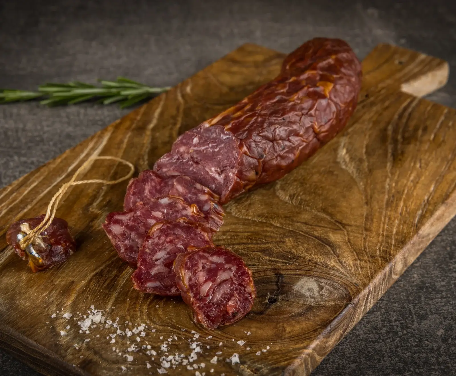 Eine angeschnittene, dunkelrote Rohwurst auf einem rustikalen Holzschneidebrett, umgeben von grobem Meersalz und einem Rosmarinzweig auf dunklem Hintergrund.