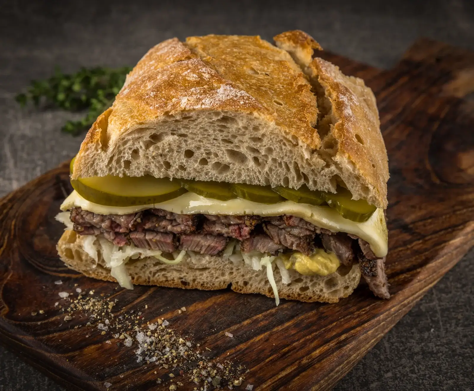 Ein großes, aufgeschnittenes Sandwich mit knusprigem Brot, gefüllt mit zartem Rindfleisch, geschmolzenem Käse, sauren Gurken, Krautsalat und Senf, appetitlich auf einem Holzbrett angerichtet.