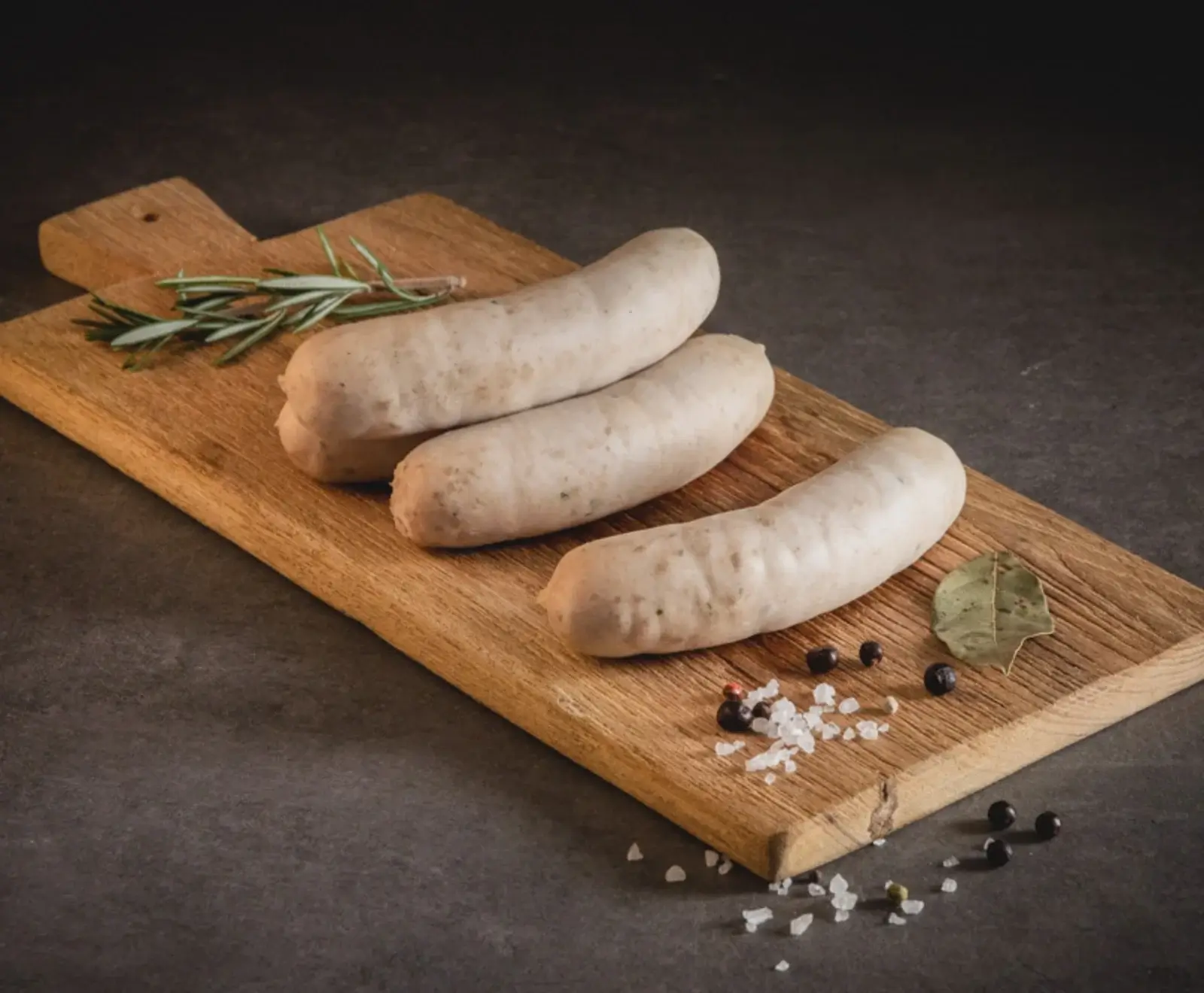 Drei rohe, helle Würste liegen diagonal auf einem Holzschneidebrett, umgeben von Rosmarin, einem Lorbeerblatt, grobem Meersalz und schwarzen Pfefferkörnern auf dunklem Untergrund.
