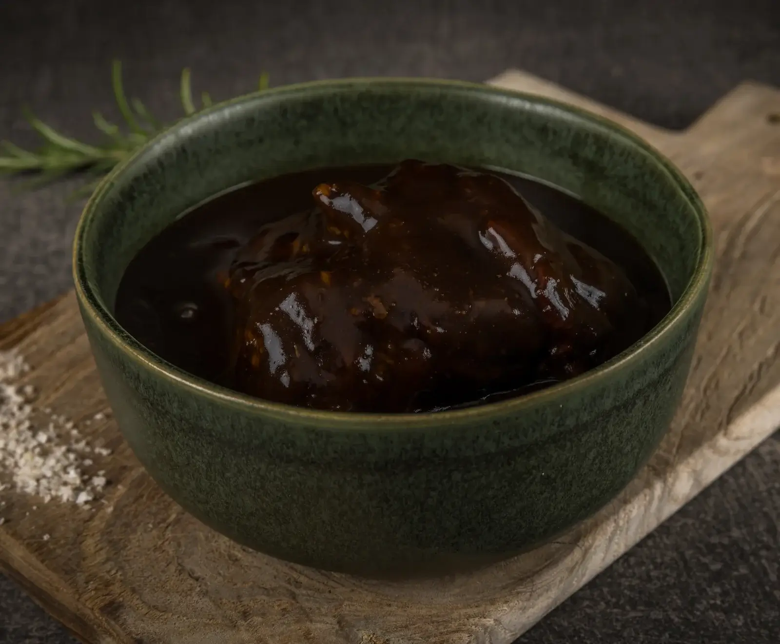 Dunkelbraune, glänzende, stückige Sauce in einer dunkelgrünen Keramikschale auf einem rustikalen Holzbrett, mit Rosmarin im unscharfen Hintergrund.