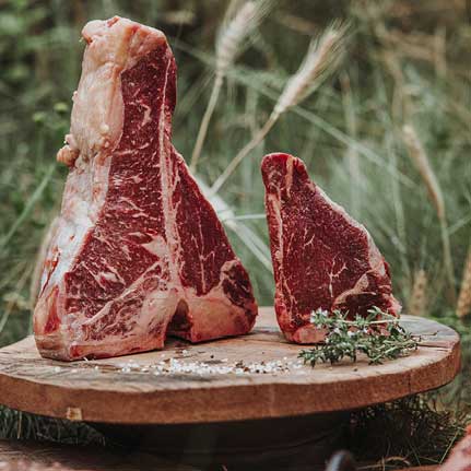 Zwei rohe, marmorierte T-Bone-Steaks stehen aufrecht auf einem rustikalen Holzbrett. Grobes Salz und frischer Thymian liegen daneben. Im unscharfen Hintergrund sind grüne Pflanzen und Getreideähren zu sehen.