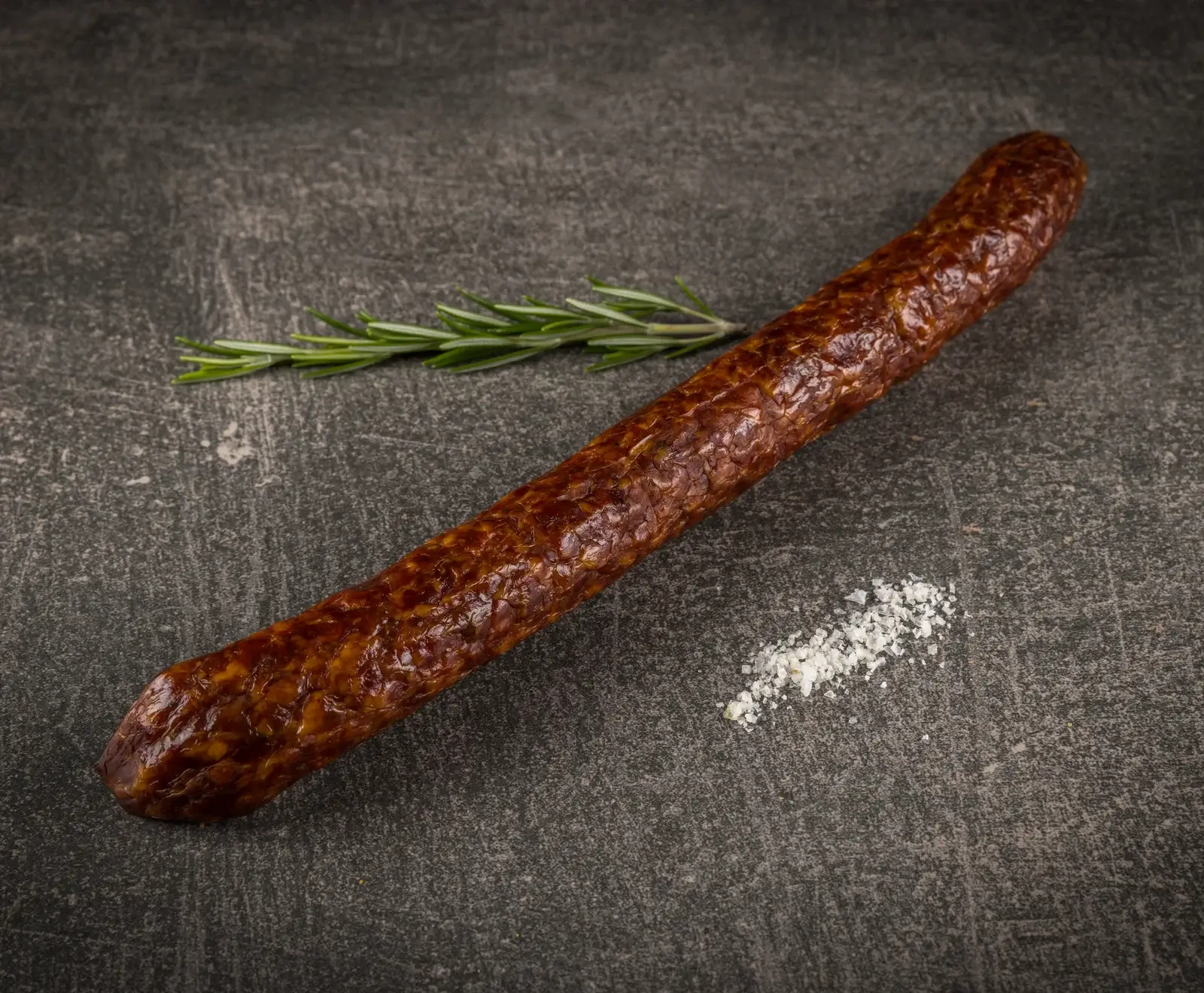 Eine lange, dunkelbraune Rohwurst liegt diagonal auf einer dunklen Steinplatte, begleitet von einem Rosmarinzweig und grobem Meersalz.
