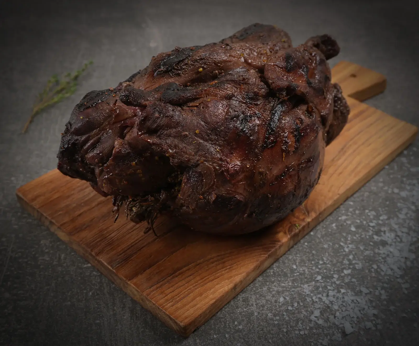 Ein großes, dunkelbraun gebratenes Stück Fleisch mit glänzender, leicht verkohlter Kruste und Gewürzen liegt auf einem rustikalen Holzschneidebrett. Im Hintergrund sind ein Thymianzweig und grobes Salz auf dunklem Untergrund zu sehen.