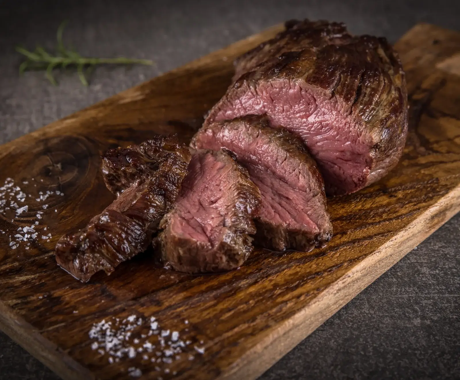 Ein aufgeschnittenes, medium-rare gebratenes Stück Rindfleisch auf einem Holzbrett, bestreut mit grobem Salz und einem Rosmarinzweig im Hintergrund.