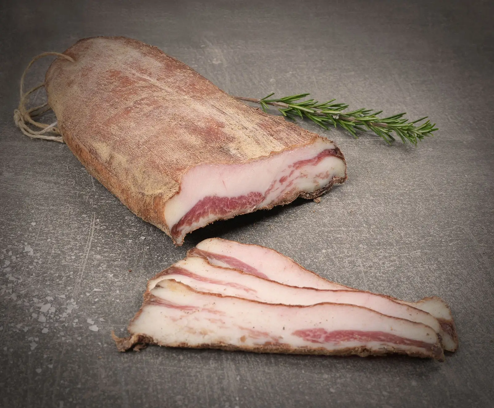 Ein großes, angeschnittenes Stück Pancetta (gepökelter Schweinebauch) mit sichtbaren Fett- und Fleischschichten liegt auf dunklem Untergrund. Daneben sind mehrere dünne Scheiben aufgefächert. Ein Rosmarinzweig ist im Hintergrund platziert.