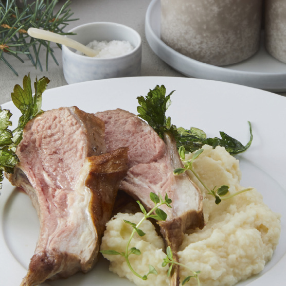 Zwei rosa gebratene Lammkoteletts mit cremigem Kartoffelpüree, garniert mit frischen Kräutern, auf einem weißen Teller. Im Hintergrund sind ein Salzschälchen und Tannenzweige angedeutet.