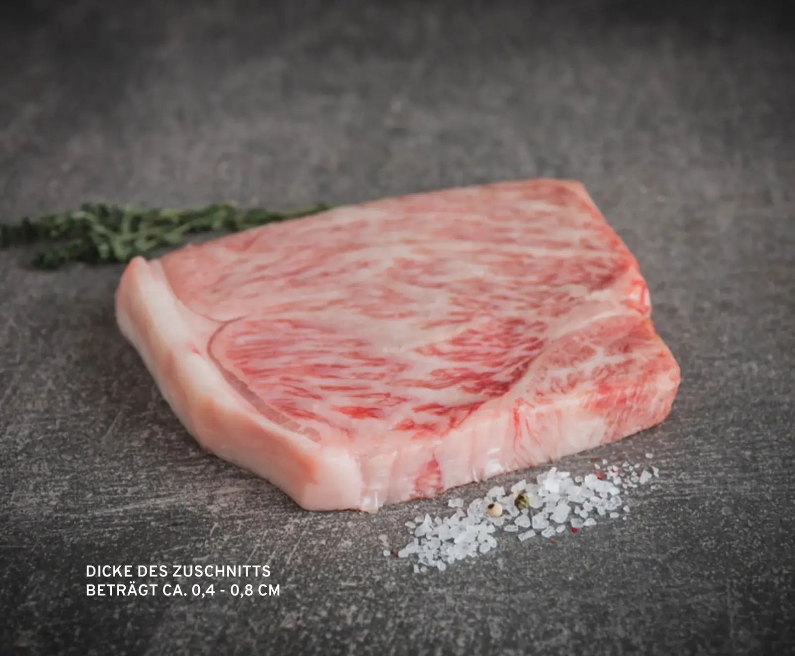 Ein rohes, stark marmoriertes Stück Rindfleisch mit Fettrand liegt auf einem dunklen Untergrund. Daneben sind grobes Salz und Pfefferkörner zu sehen, im Hintergrund ein Kräuterzweig.