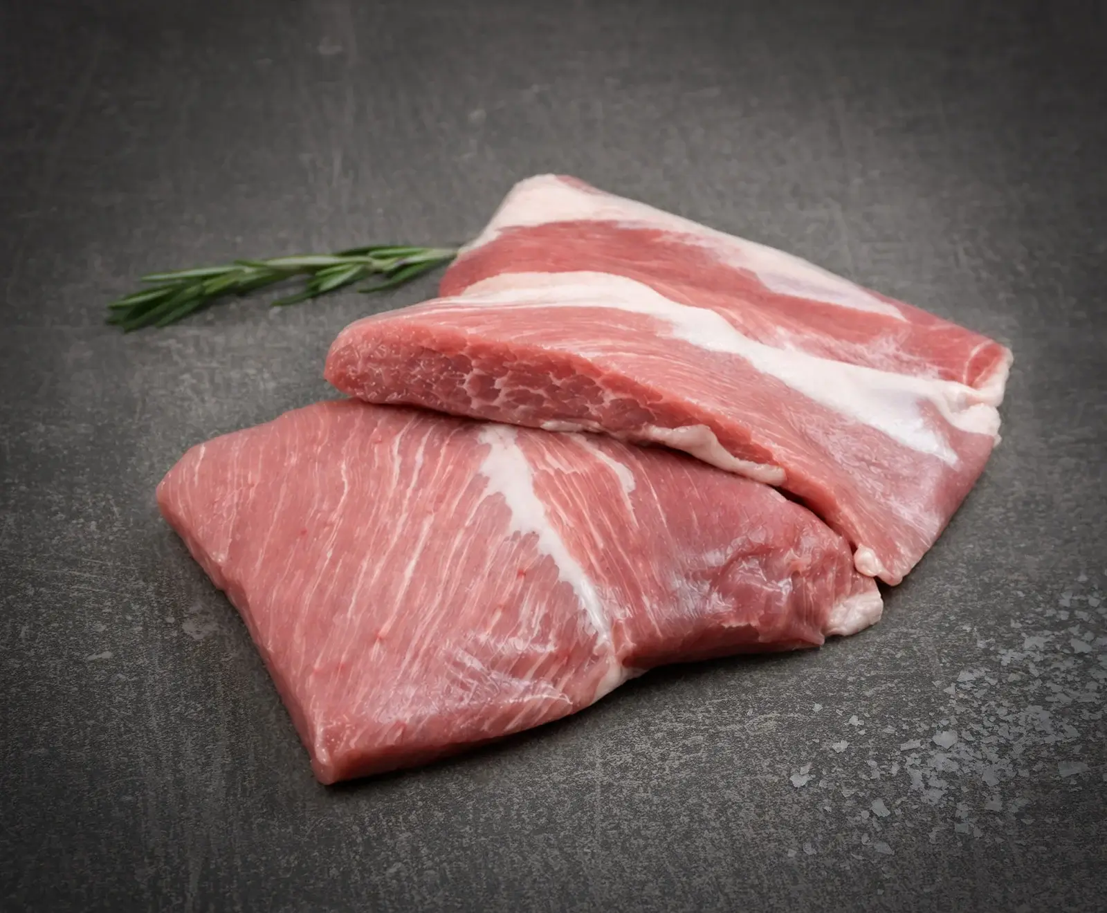 Zwei Stücke roher Schweinebauch, übereinanderliegend, mit sichtbaren Fett- und Muskelschichten. Ein Rosmarinzweig liegt daneben auf einem dunklen, strukturierten Untergrund.