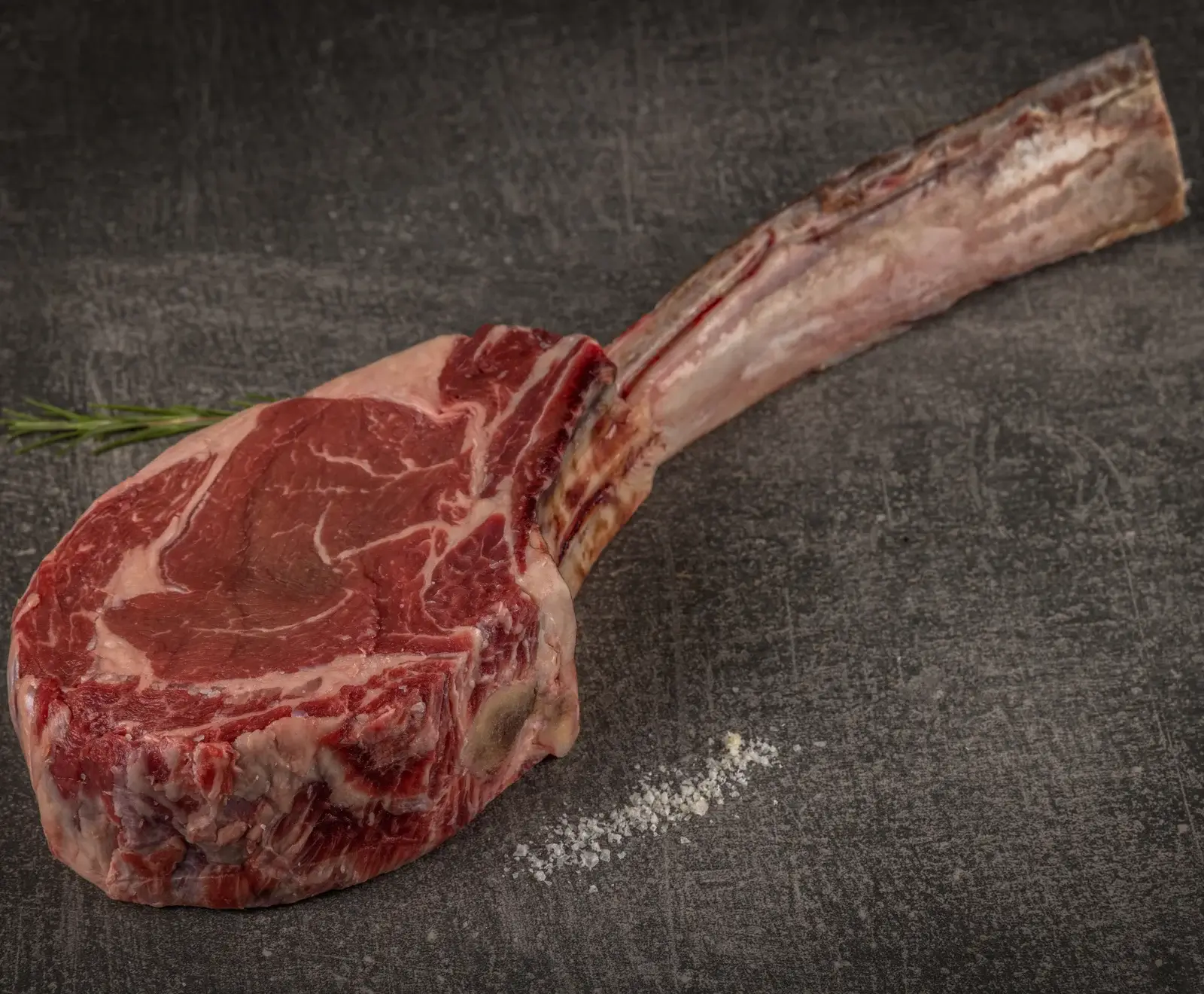Ein rohes Tomahawk-Steak mit langem Knochen liegt auf einer dunklen, strukturierten Oberfläche. Das rote Fleisch zeigt deutliche Marmorierung. Links oben ist ein Rosmarinzweig, rechts unten grobes Salz.