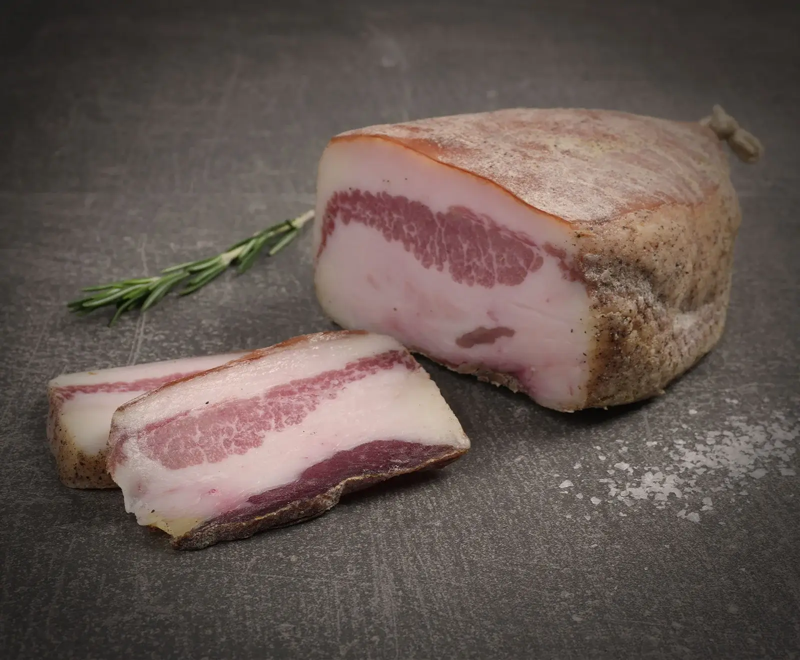 Ein großes Stück geräucherter Bauchspeck (Pancetta) mit zwei geschnittenen Scheiben auf dunklem Untergrund, garniert mit Rosmarin und grobem Salz.
