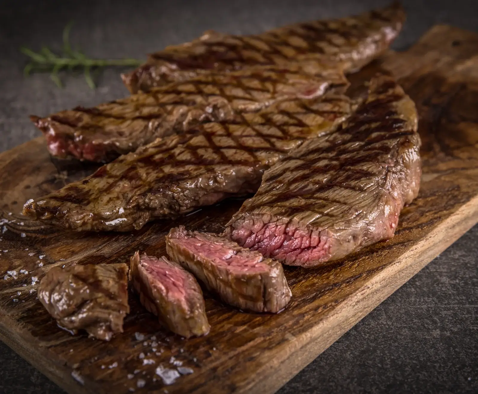 Gegrilltes Steak auf einem Holzbrett, teilweise in Scheiben geschnitten, mit rosa Kern. Grillspuren sind sichtbar, grobes Salz und ein Rosmarinzweig liegen daneben.