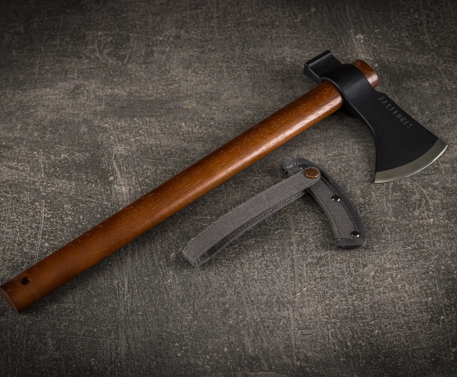 Ein Barebones Beil mit braunem Holzgriff und schwarzem Axtkopf mit scharfer Klinge liegt auf dunklem, strukturiertem Untergrund. Daneben eine graue Stoffscheide.