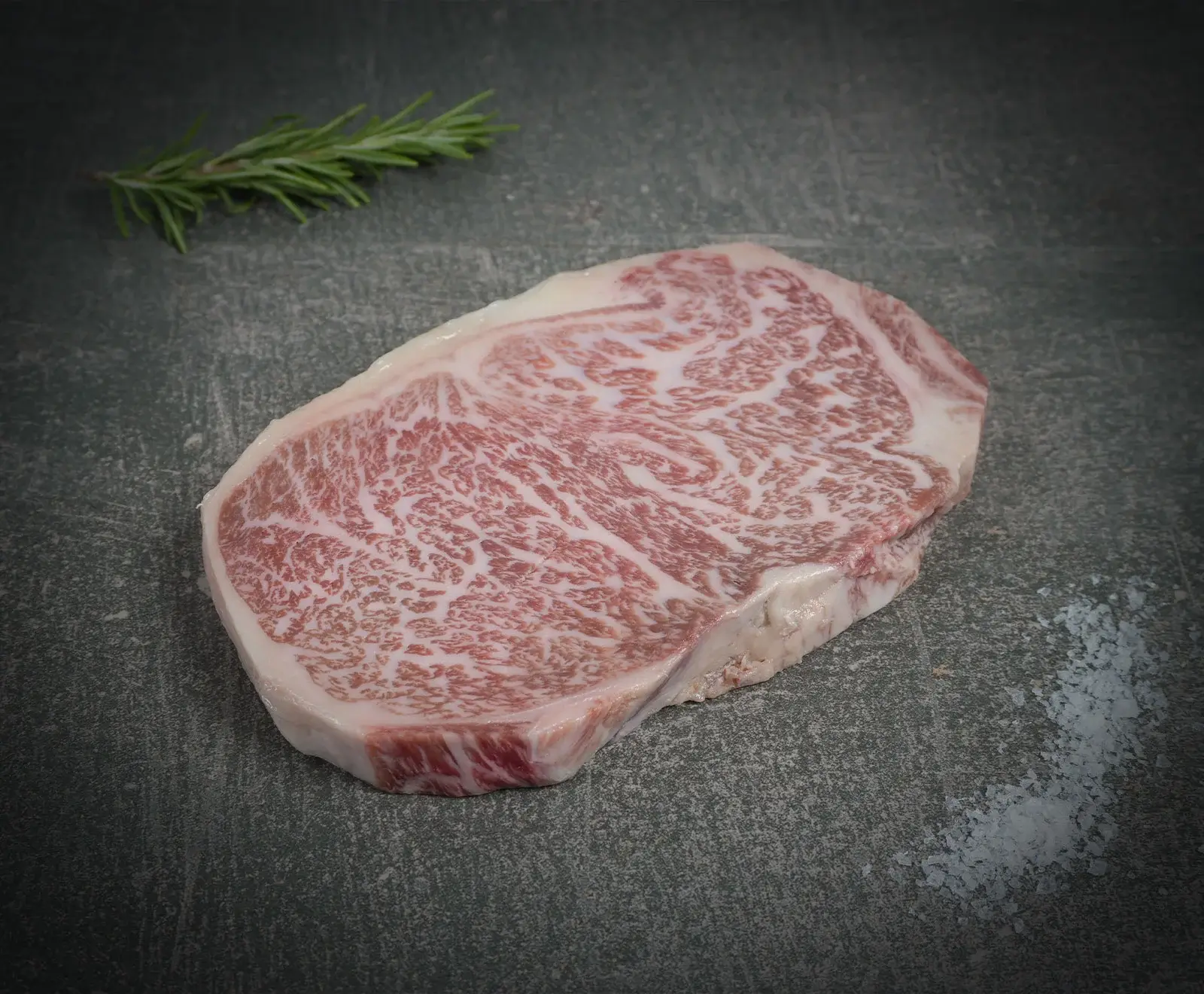 Nahaufnahme eines rohen, stark marmorierten Rindersteaks auf dunklem Untergrund, mit Rosmarin und Meersalz.