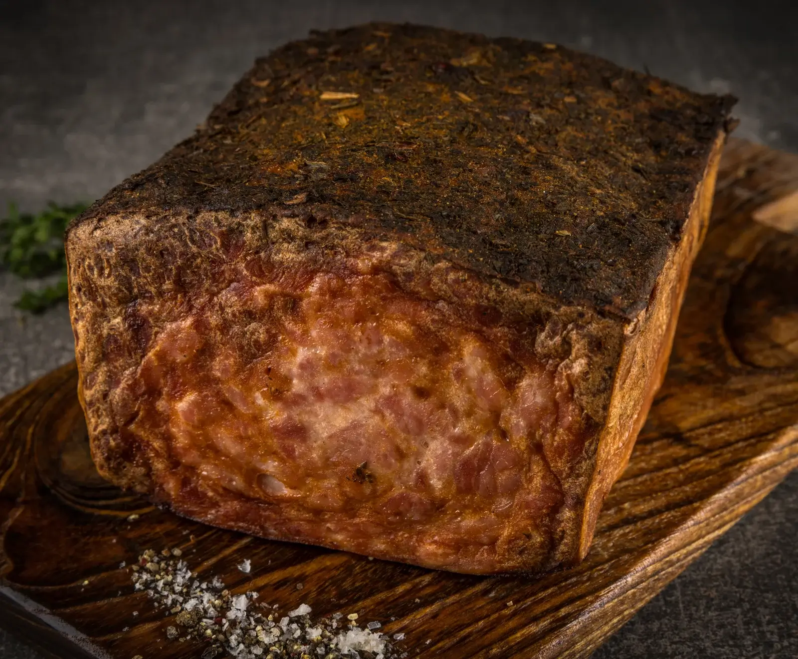 Ein großes, rechteckiges Stück gepökeltes oder geräuchertes Fleisch mit dunkler, würziger Kräuterkruste und rötlich-rosa, marmoriertem Inneren, auf einem Holzbrett mit grobem Salz und Pfeffer.