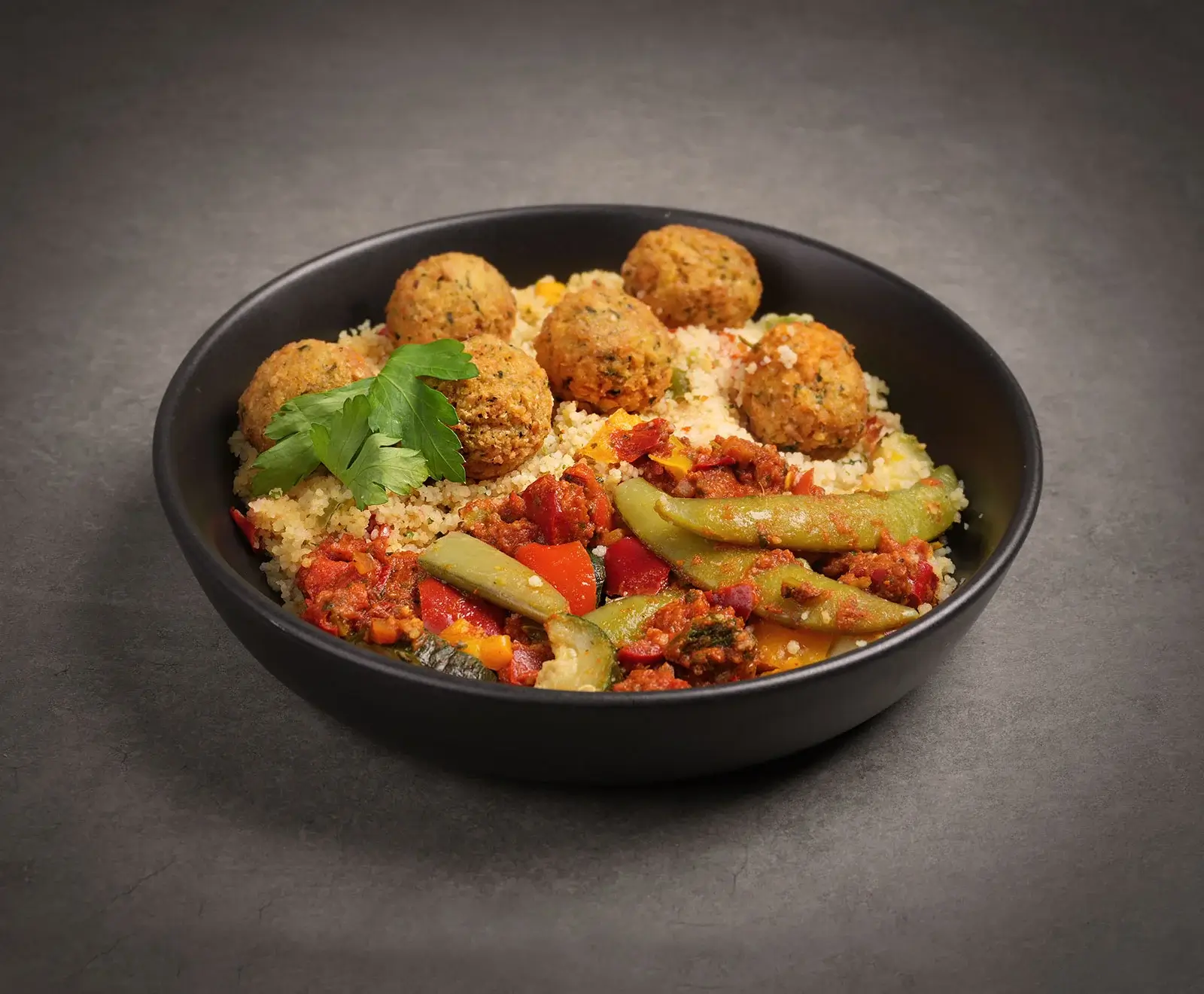 Eine schwarze Schale gefüllt mit Couscous, goldbraunen Falafelbällchen, gebratenem Gemüse (rote Paprika, grüne Zuckerschoten, Zucchini) und frischer Petersilie, auf dunklem Untergrund.