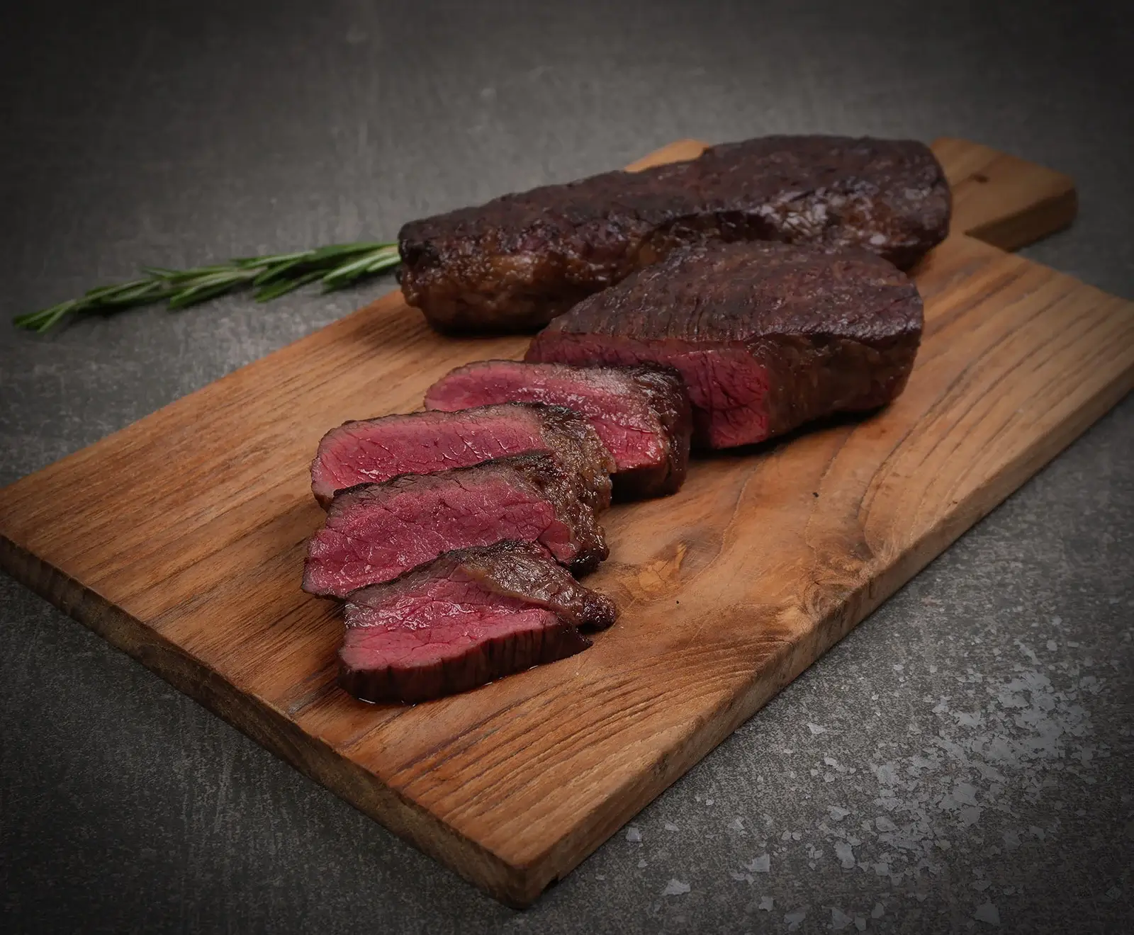 Ein aufgeschnittenes, medium-rare gegartes Steak auf einem Holzschneidebrett, daneben ein weiteres Stück Fleisch und ein Rosmarinzweig. Der Hintergrund ist dunkelgrau mit Salzflocken.