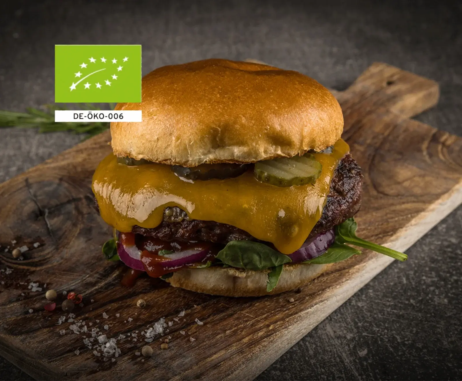 Ein appetitlicher Bio-Burger mit geschmolzenem Käse, Rindfleisch-Patty, Gurken, roten Zwiebeln und Salat auf einem rustikalen Holzbrett. Grobes Salz und Pfeffer sind verstreut. Das EU-Bio-Logo ist oben links sichtbar.