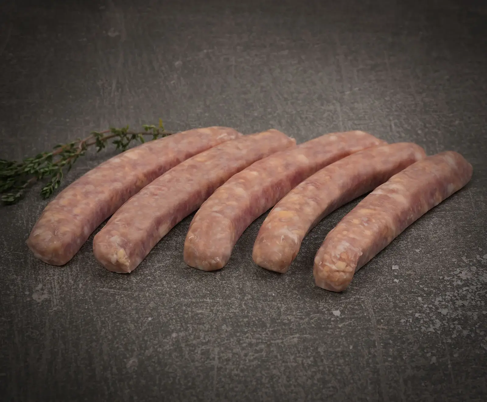 Fünf rohe, hellrosa Würste liegen nebeneinander auf einer dunklen, strukturierten Oberfläche. Ein kleiner Thymianzweig ist links oben zu sehen.