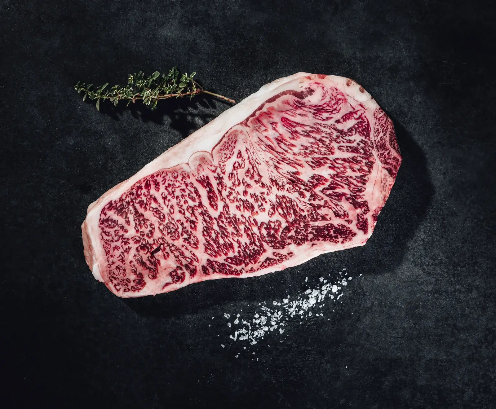 Ein rohes, stark marmoriertes Wagyu-Steak liegt auf einer dunklen Oberfläche, begleitet von einem Thymianzweig und grobem Meersalz. Das Fleisch zeigt eine intensive Fettmaserung.
