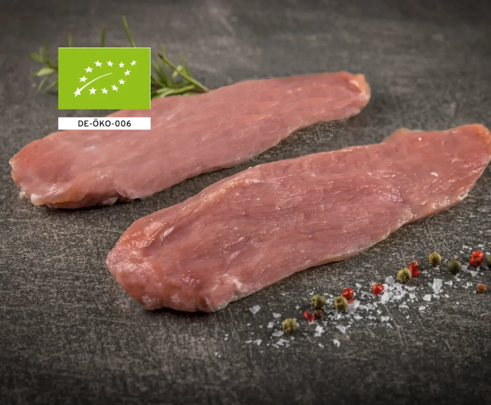 Nahaufnahme von zwei Bio-Schweinefilets auf einer dunklen Schieferplatte, garniert mit Rosmarin, Meersalz und bunten Pfefferkörnern. Das EU-Bio-Logo DE-ÖKO-006 ist sichtbar.