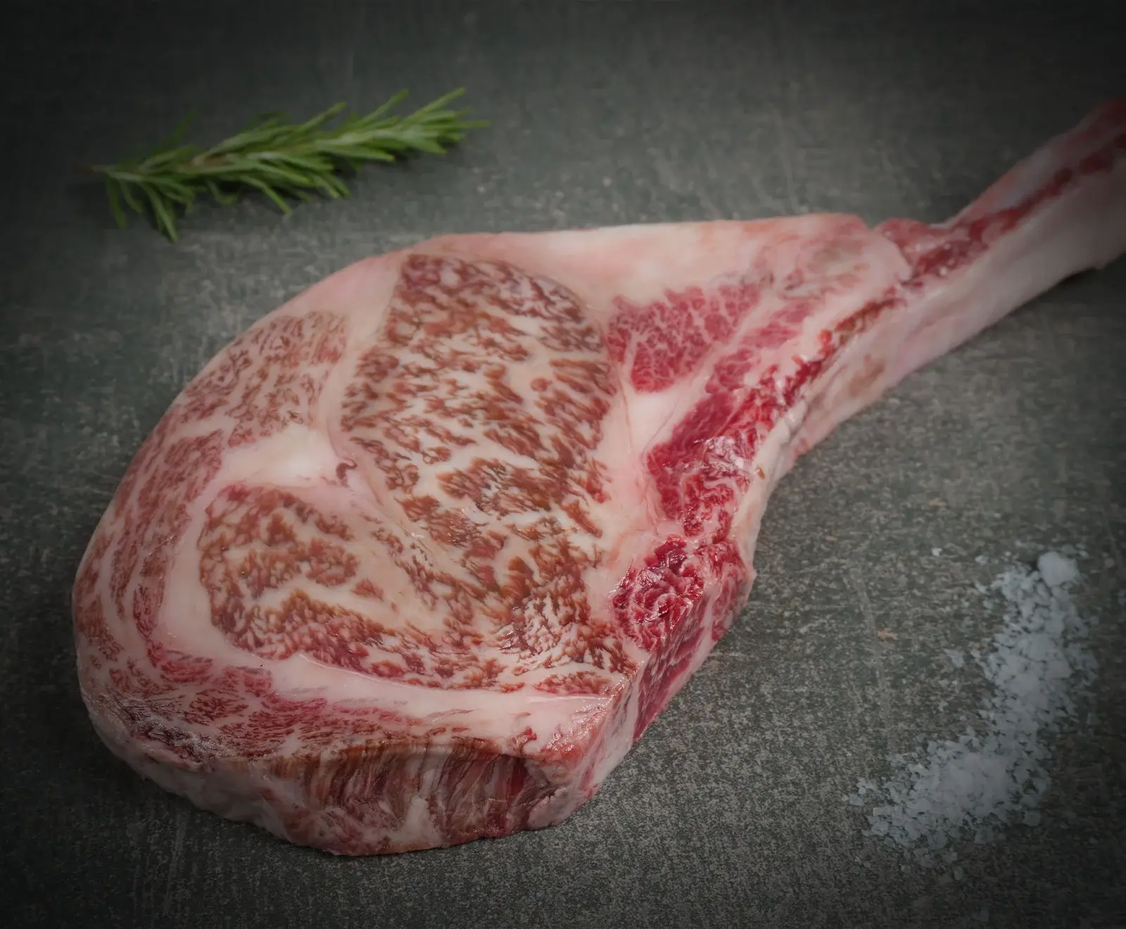 Ein rohes Tomahawk-Steak mit ausgeprägter Marmorierung liegt auf einer dunklen, strukturierten Oberfläche. Links oben ist ein Rosmarinzweig zu sehen, rechts unten grobes Meersalz.