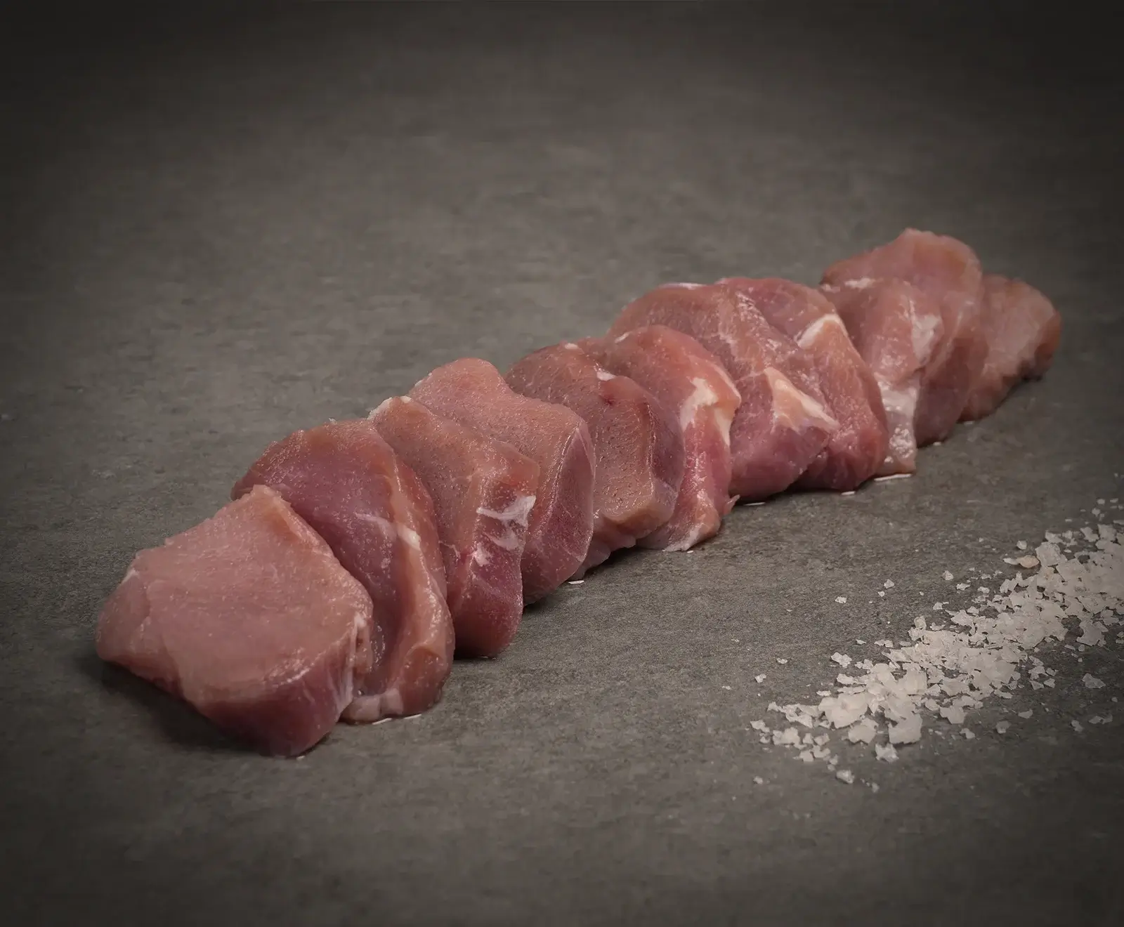 Rohe Schweinefiletscheiben liegen auf einer dunklen, steinernen Oberfläche, daneben eine kleine Menge grobes Meersalz. Die rosa Fleischstücke sind in einer Reihe angeordnet.