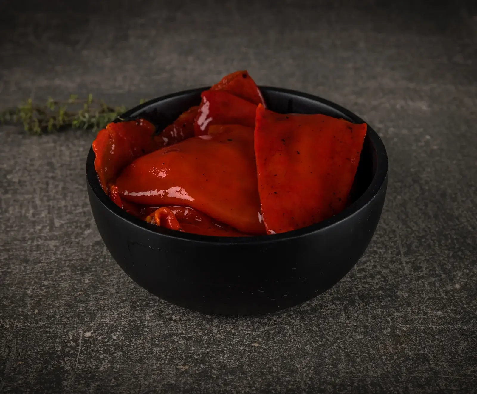 Eine schwarze Schale gefüllt mit glänzenden, roten Paprikastücken, die mariniert oder geröstet aussehen, auf einem dunklen, strukturierten Hintergrund mit einem Thymianzweig.
