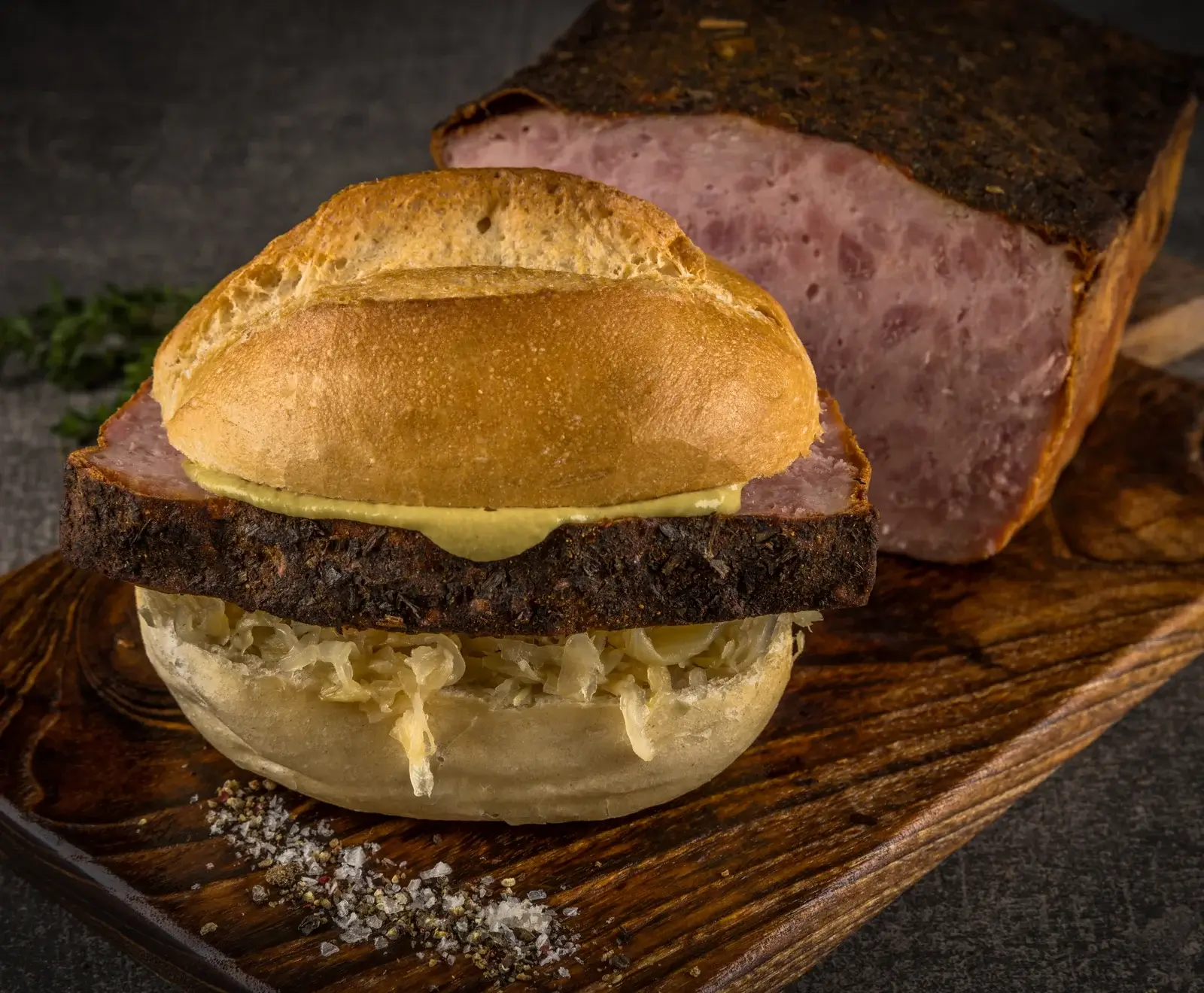Ein Leberkäse-Brötchen mit einer dicken Scheibe Leberkäse, Senf und Sauerkraut auf einem rustikalen Holzbrett. Im Hintergrund ist ein angeschnittener Leberkäse-Laib zu sehen. Salz und Pfeffer sind auf dem Brett verstreut.