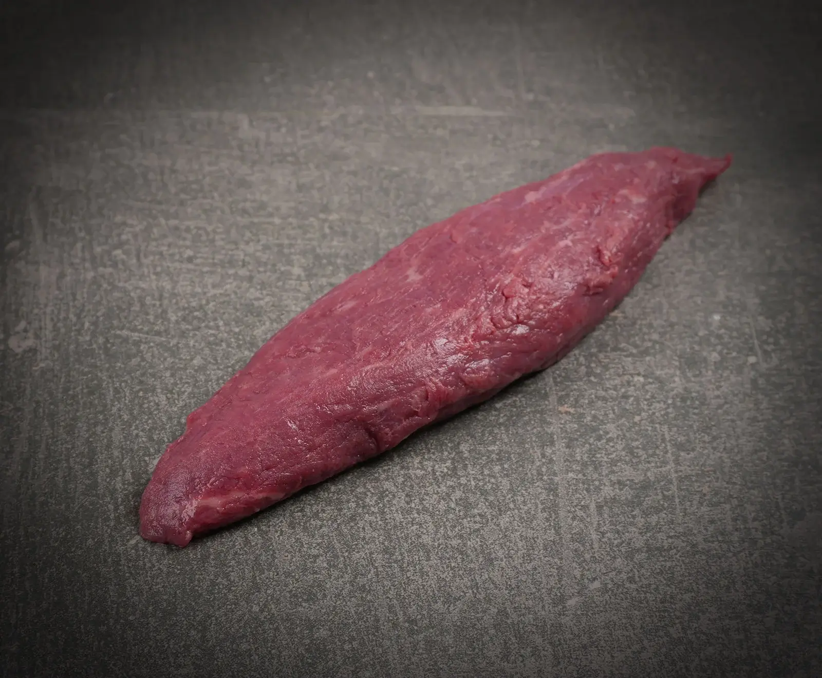 Ein längliches Stück rohes, dunkelrotes Fleisch, vermutlich Filet oder Lende, liegt diagonal auf einer dunklen, strukturierten Oberfläche.
