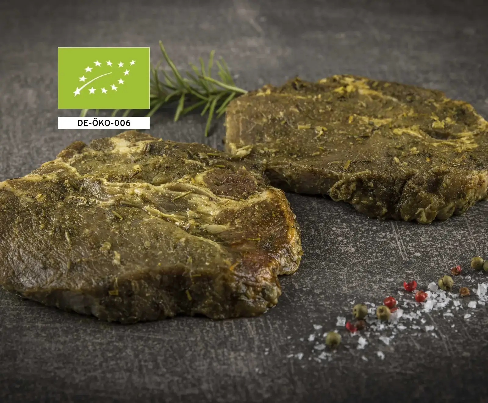Zwei kräutermarinierte Bio-Schweinenackensteaks auf dunklem Untergrund, mit Rosmarin, Salz und Pfeffer. Links oben das EU-Bio-Siegel DE-ÖKO-006.