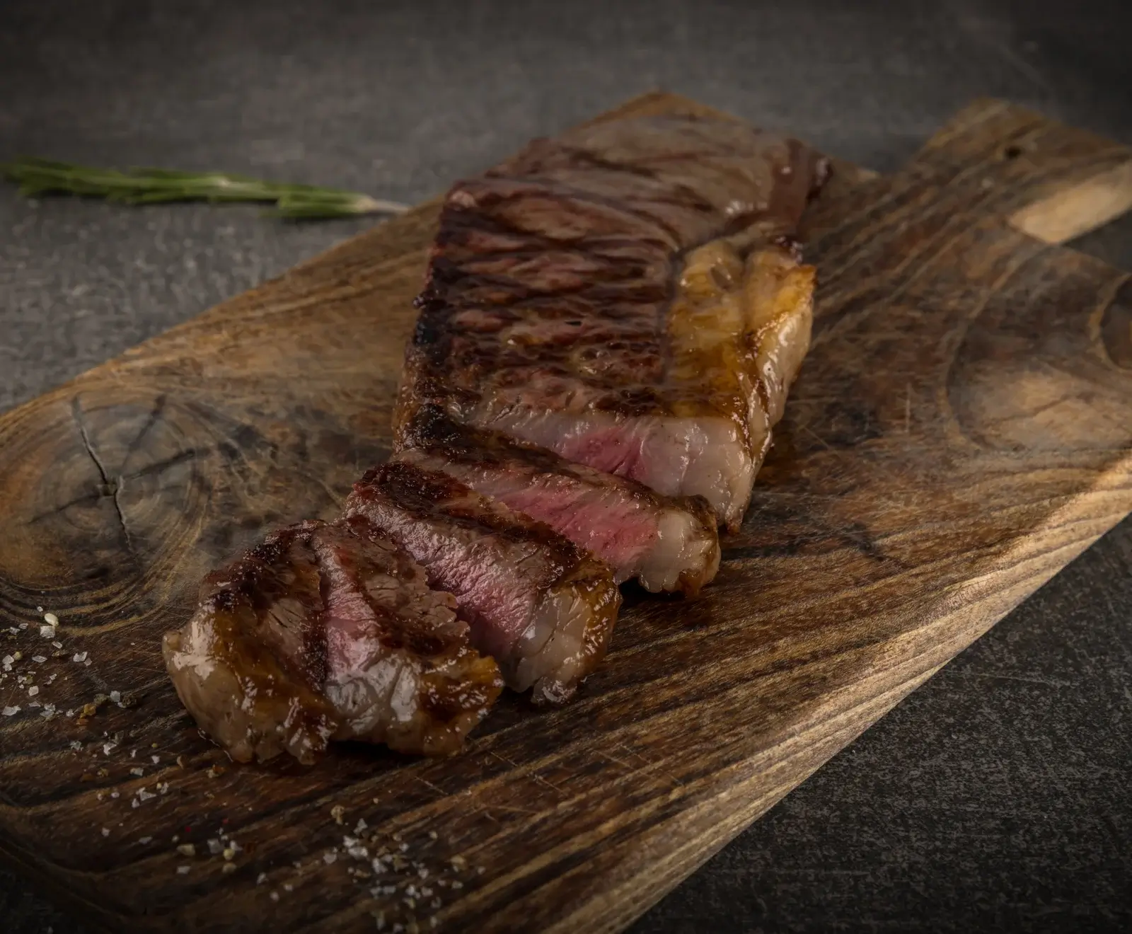 Ein perfekt gegrilltes Rindersteak, teilweise in Scheiben geschnitten, mit rosa Kern, liegt auf einem rustikalen Holzbrett. Grobes Salz und ein Rosmarinzweig sind im Hintergrund zu sehen.
