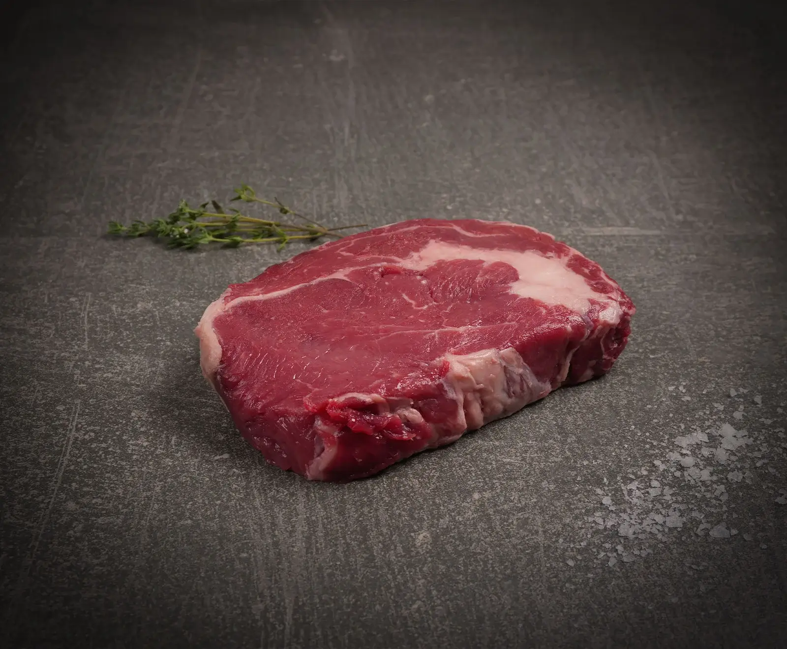 Ein rohes Ribeye-Steak mit deutlicher Marmorierung liegt auf einer dunklen, strukturierten Oberfläche. Links daneben liegen frische Thymianzweige, rechts sind grobe Salzkristalle verstreut.