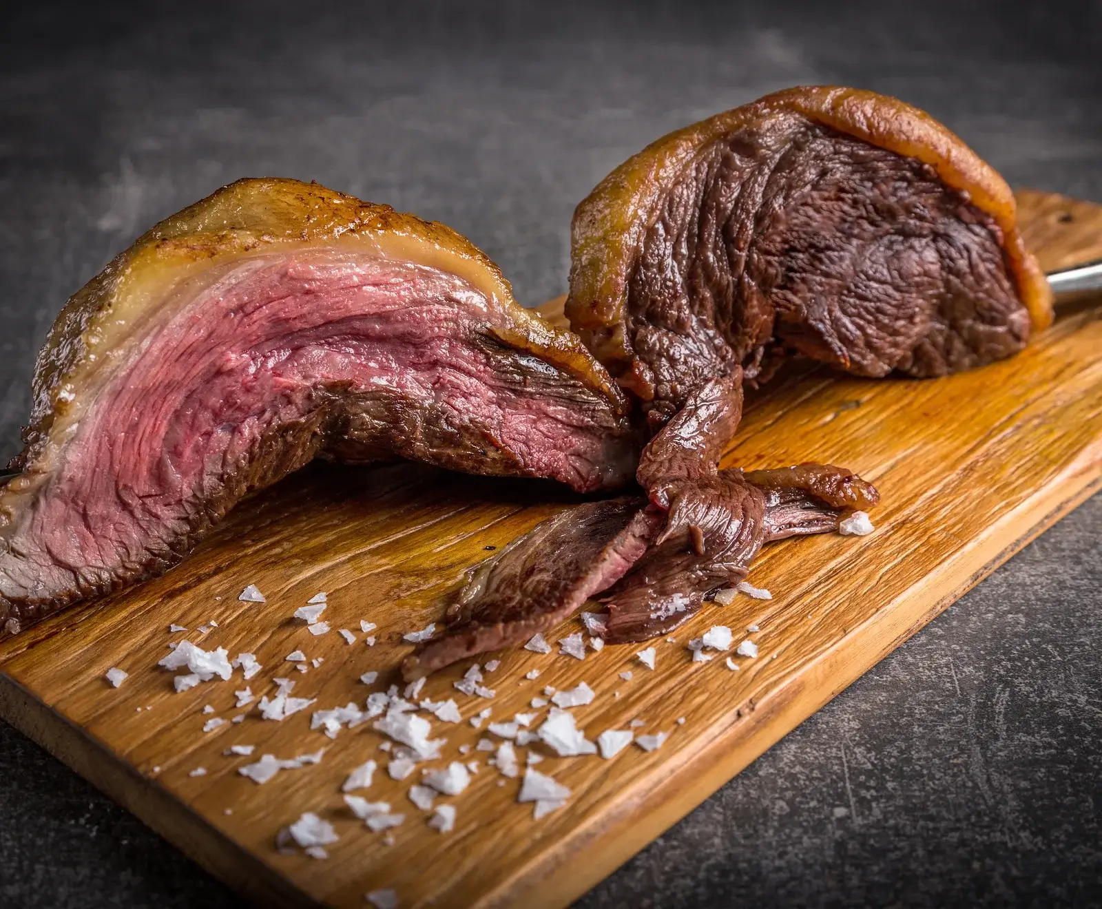 Ein aufgeschnittenes, medium-rare gegrilltes Stück Fleisch (vermutlich Picanha) mit goldbrauner Kruste und Fettschicht, auf einem Holzbrett mit grobem Meersalz.