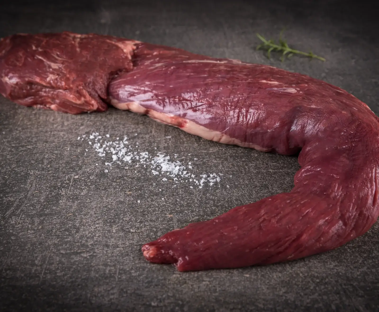 Ein rohes, dunkelrotes Rinderfilet liegt auf einer dunklen Steinplatte, daneben grobes Meersalz und ein Rosmarinzweig.