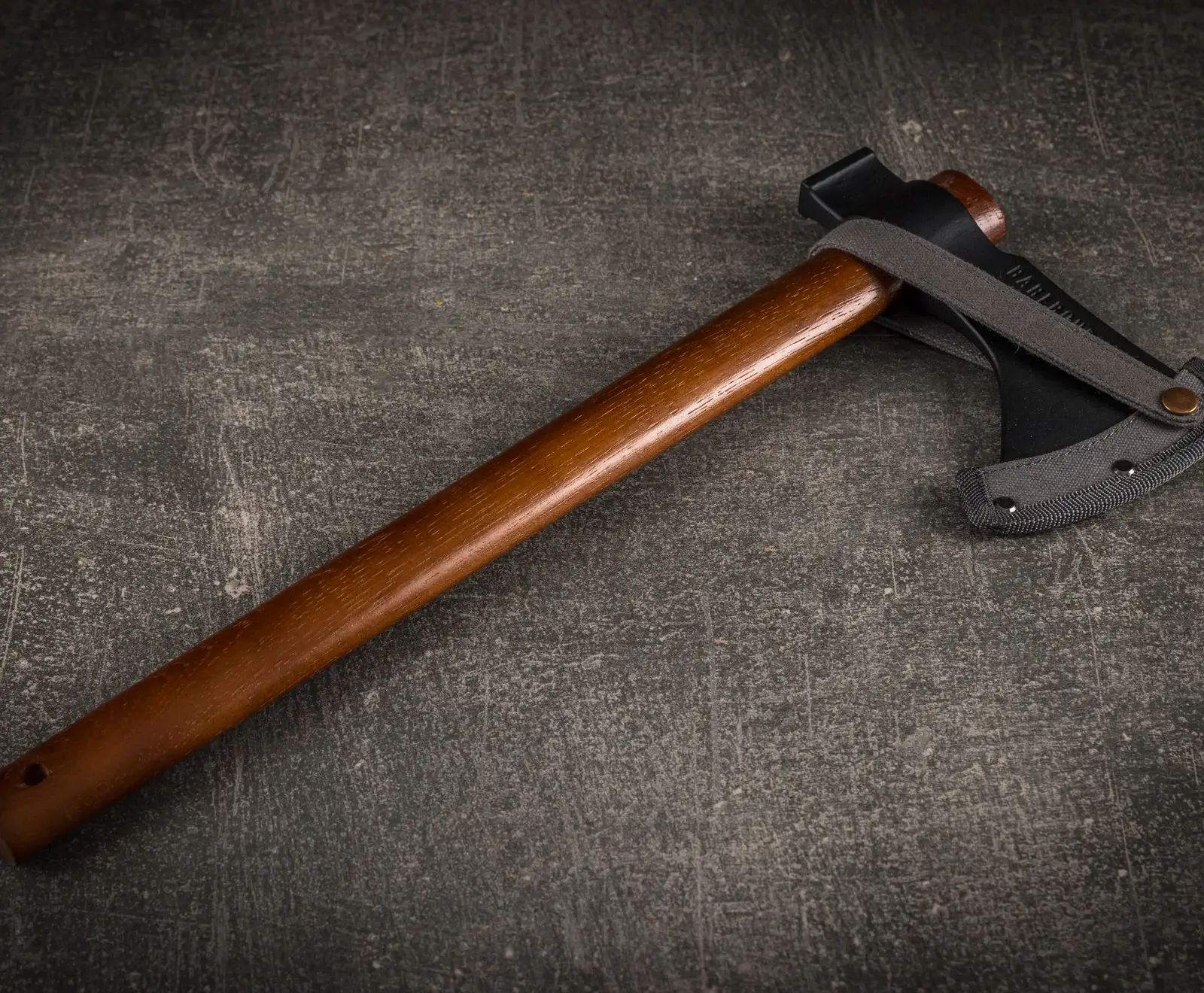 Ein kleines Beil mit braunem Holzgriff und schwarzem Axtkopf, dessen Klinge von einer grauen Stoffhülle mit Lederschlaufe geschützt wird, liegt diagonal auf dunklem, strukturiertem Untergrund.