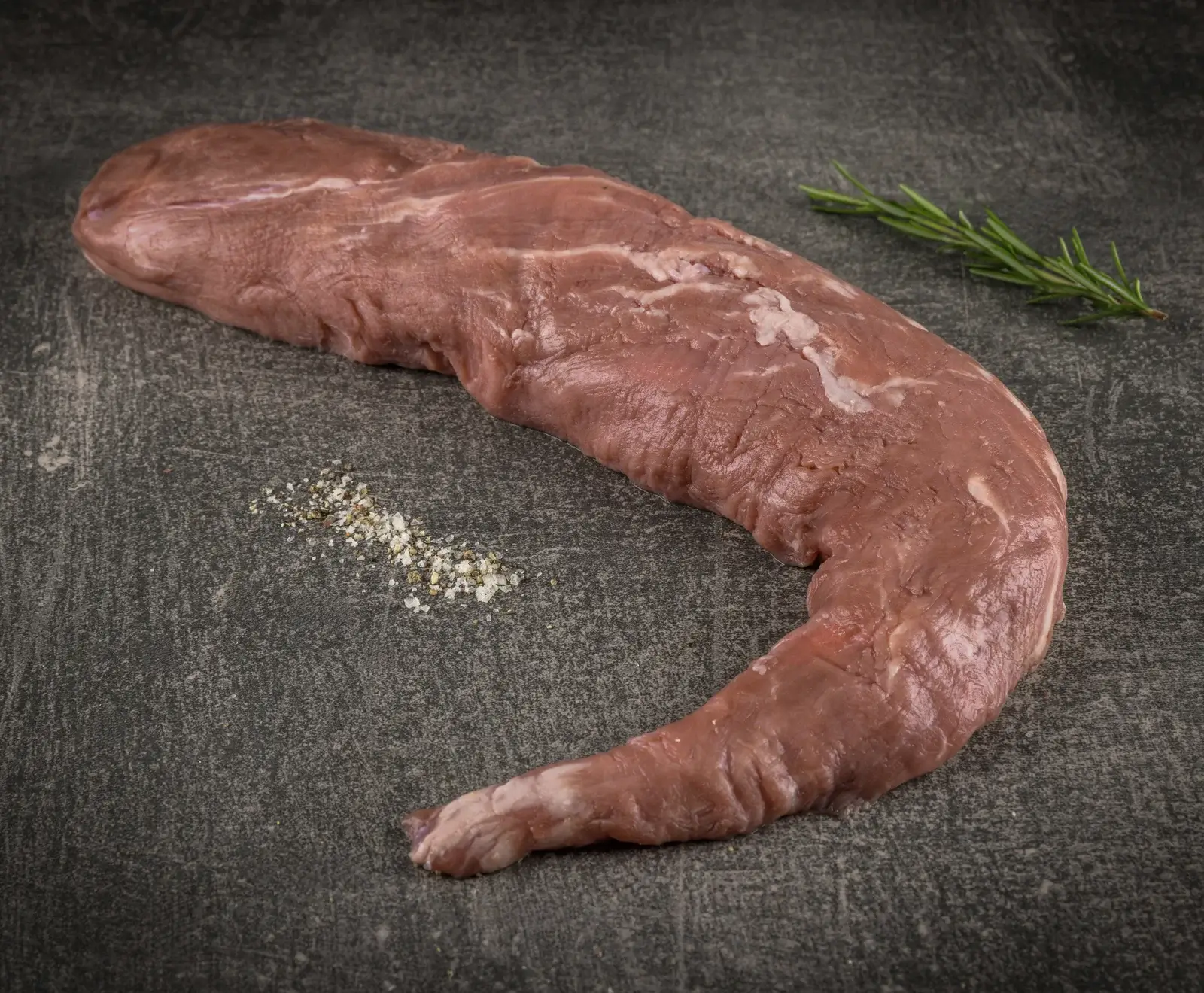 Ein rohes, zartrosa Schweinefilet liegt auf einer dunklen, strukturierten Steinplatte. Daneben befinden sich ein frischer Rosmarinzweig und eine kleine Prise grobes Salz und Pfeffer.
