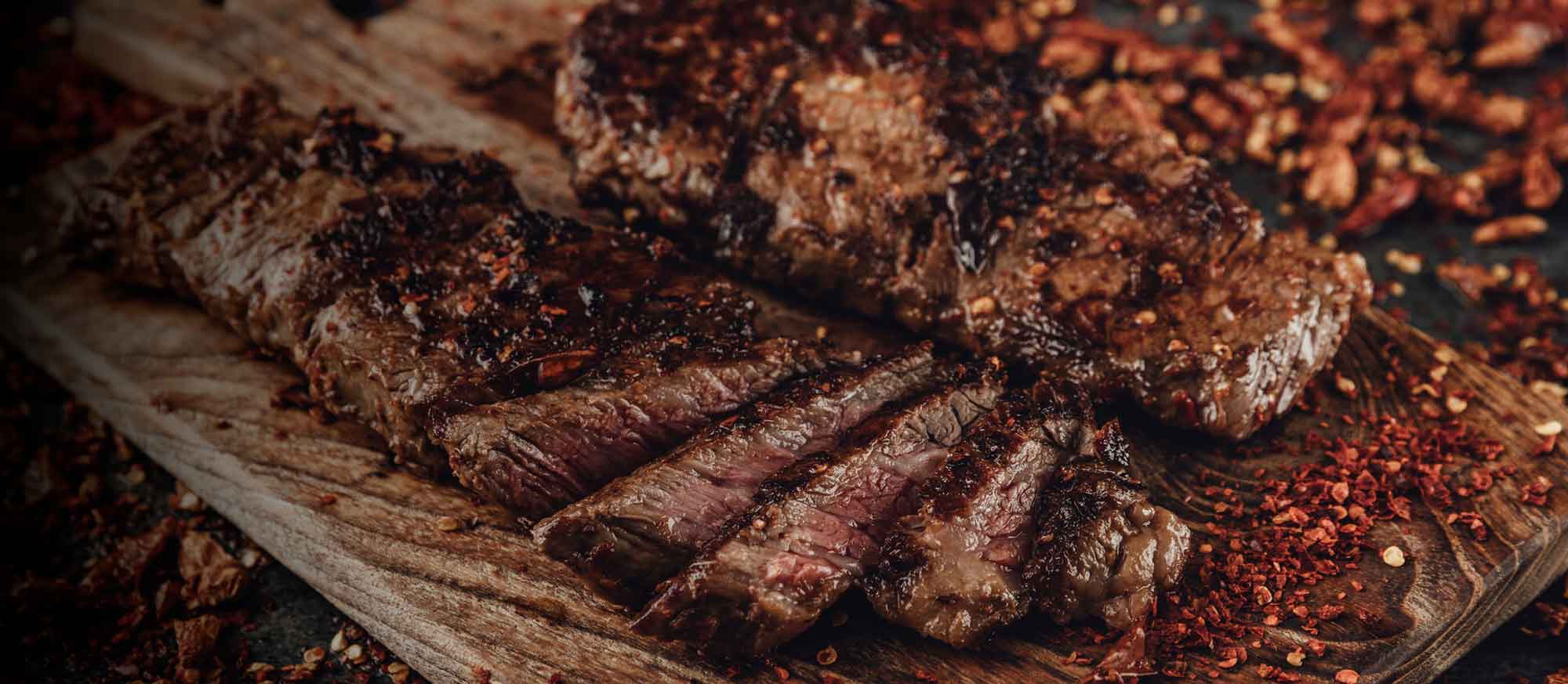 Ein saftiges, medium gegrilltes Steak, teilweise in Scheiben geschnitten, mit dunklen Gewürzen und roten Chiliflocken auf einem rustikalen Holzbrett.