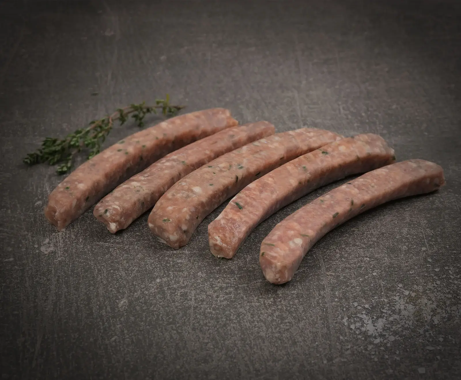 Fünf rohe, hellrosa Würste mit sichtbaren Kräutern liegen leicht gekrümmt auf einer dunklen, strukturierten Oberfläche. Ein kleiner Thymianzweig ist links oben zu sehen.