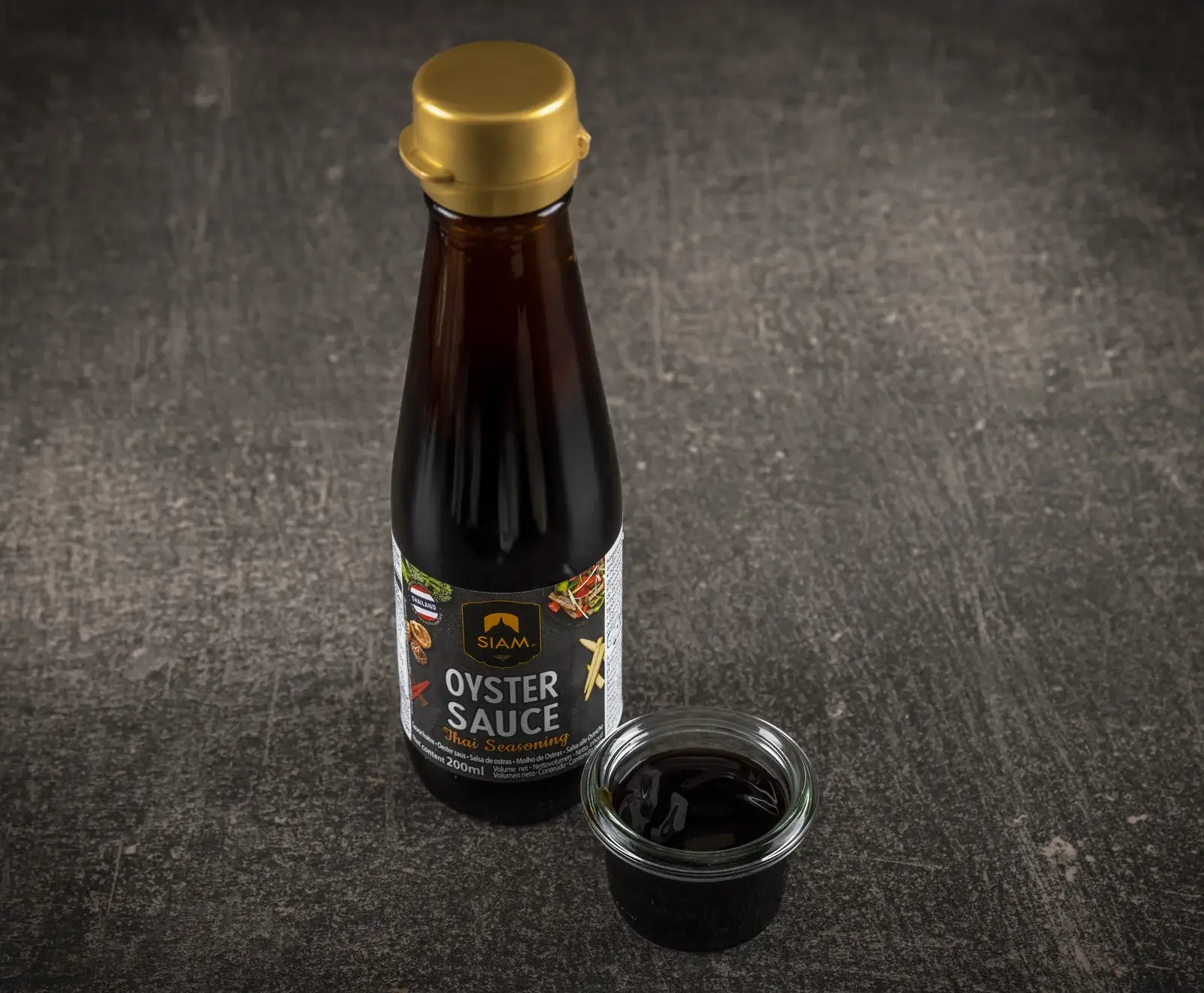Eine dunkle Glasflasche SIAM Austernsauce (Thai Seasoning, 200ml) mit goldenem Deckel steht auf einer dunklen Oberfläche. Daneben ist ein kleines Glasgefäß mit der dunklen Sauce zu sehen.