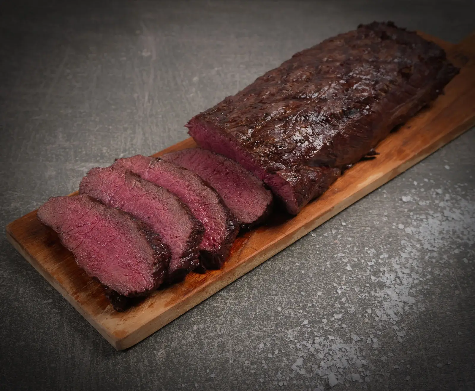 Ein saftiges, medium-rare gebratenes Fleischstück mit dunkler Kruste, teilweise in dicke Scheiben geschnitten, liegt auf einem Holzbrett. Grobes Salz ist auf dem dunklen Hintergrund verstreut.