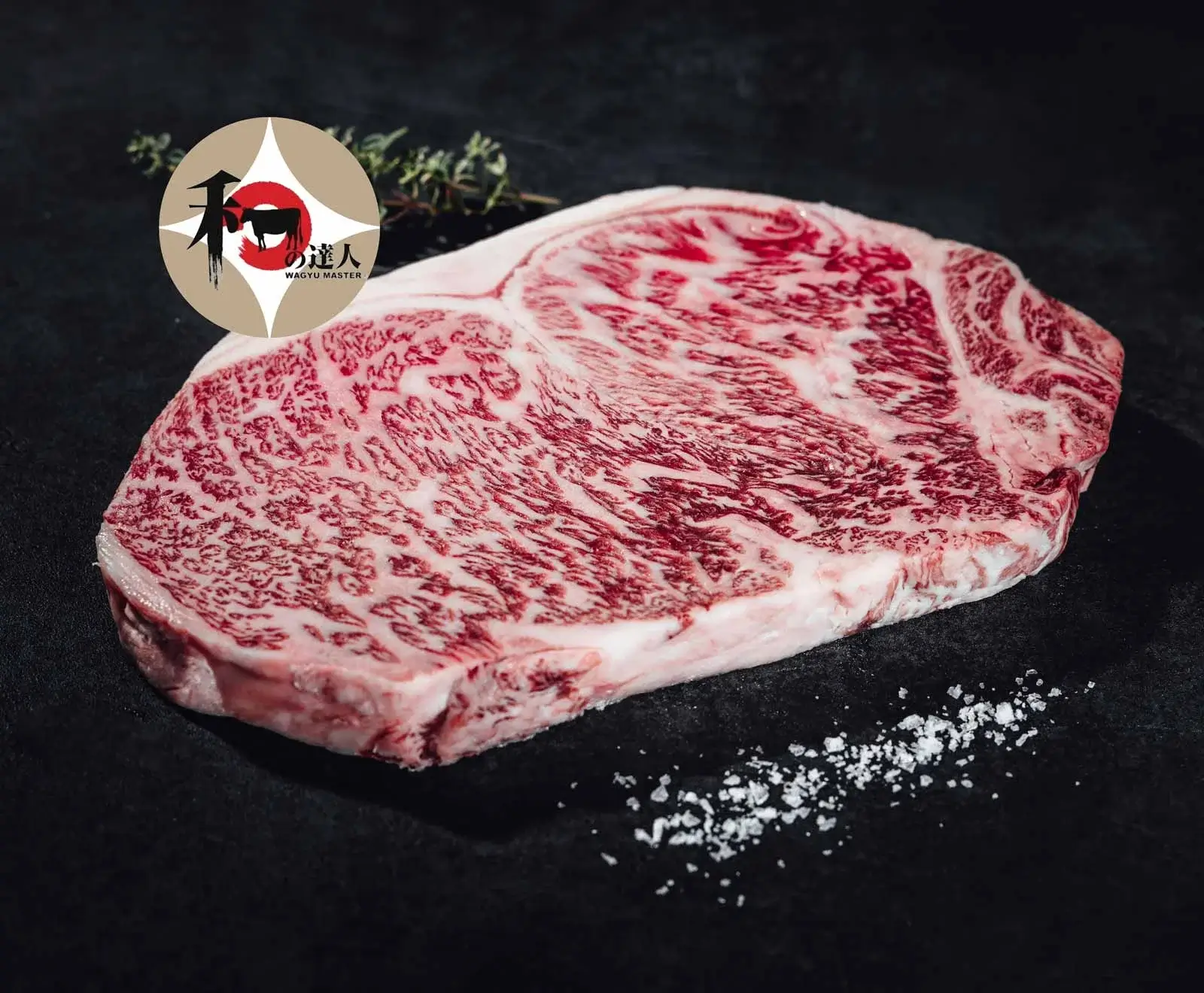 Ein rohes, stark marmoriertes Wagyu-Steak (vermutlich A5-Qualität) auf dunklem Untergrund, mit Meersalz, einem Wagyu Master Logo und Thymianzweig im Hintergrund.