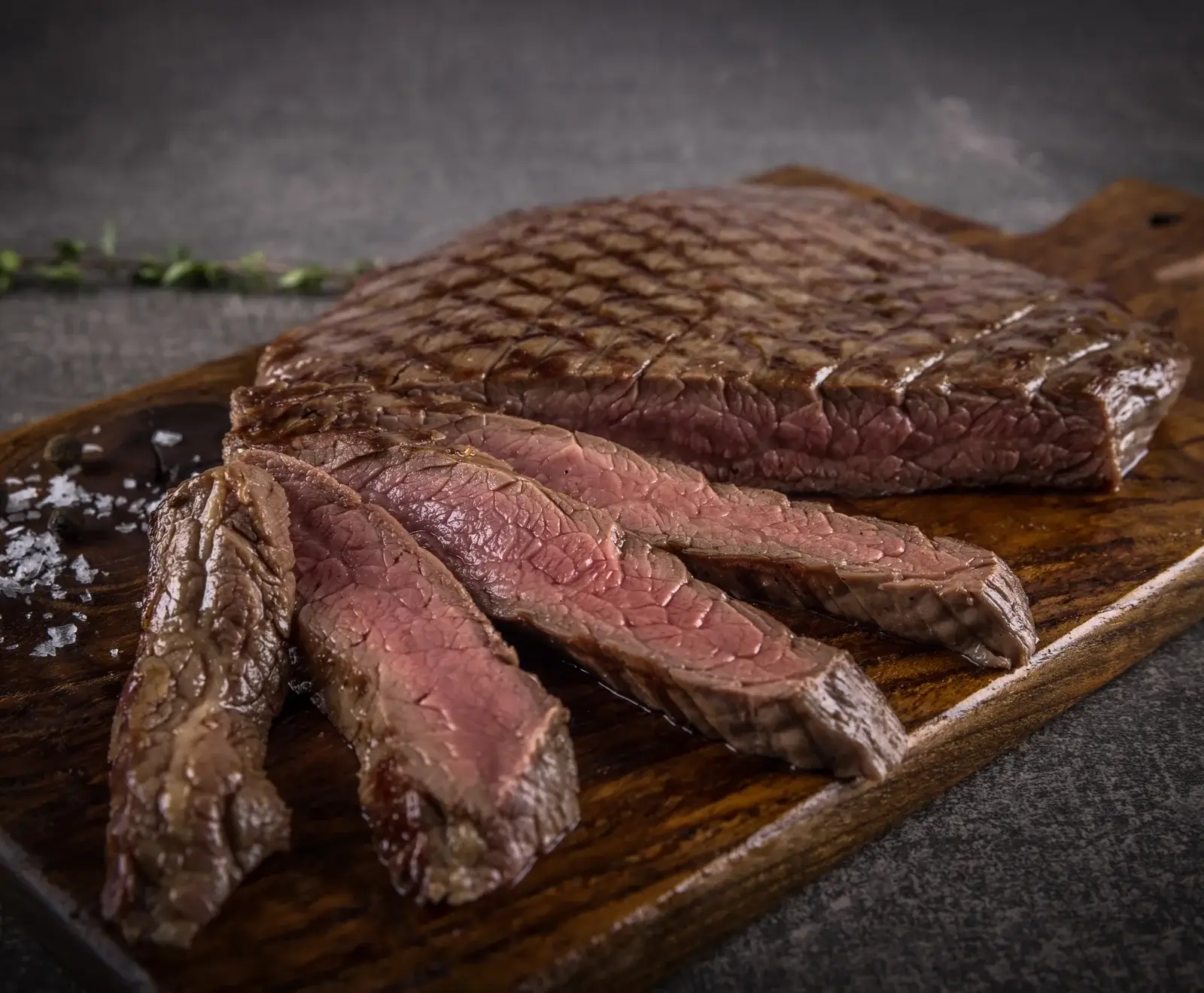 Ein perfekt gegrilltes Steak mit Grillstreifen, teilweise in saftige Scheiben geschnitten, liegt auf einem dunklen Holzbrett. Die Schnittflächen sind rosa. Grobes Salz und Pfeffer sind daneben.